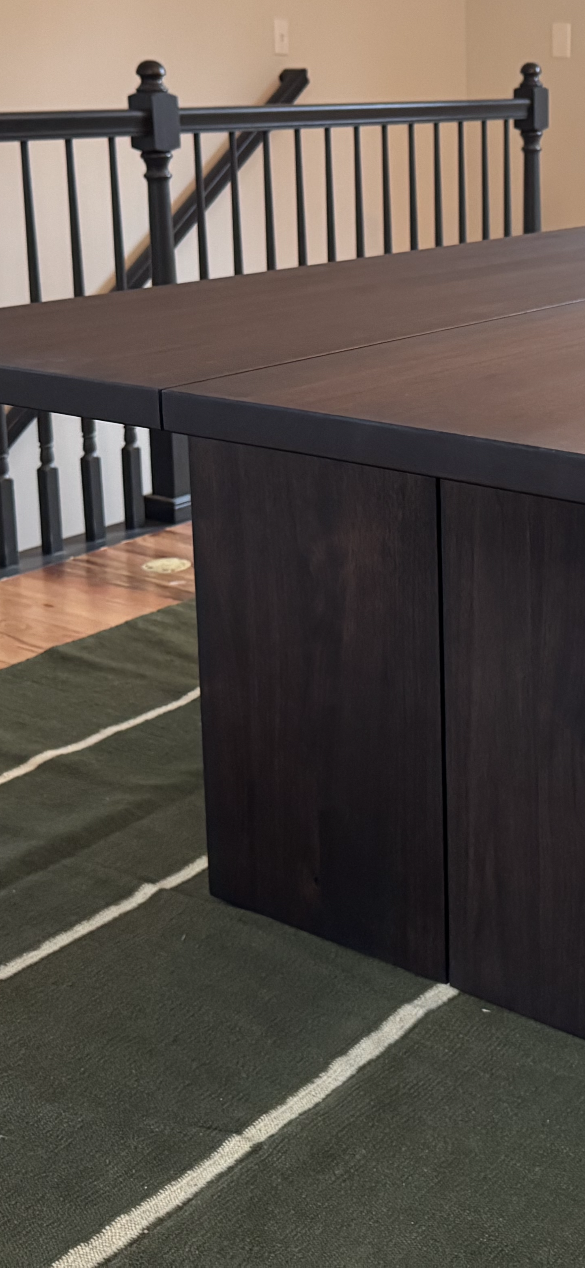 Close-up of a dark wooden dining table with a corner visible, set on a green and white striped rug. In the background, a staircase with black railing and beige wall is visible.