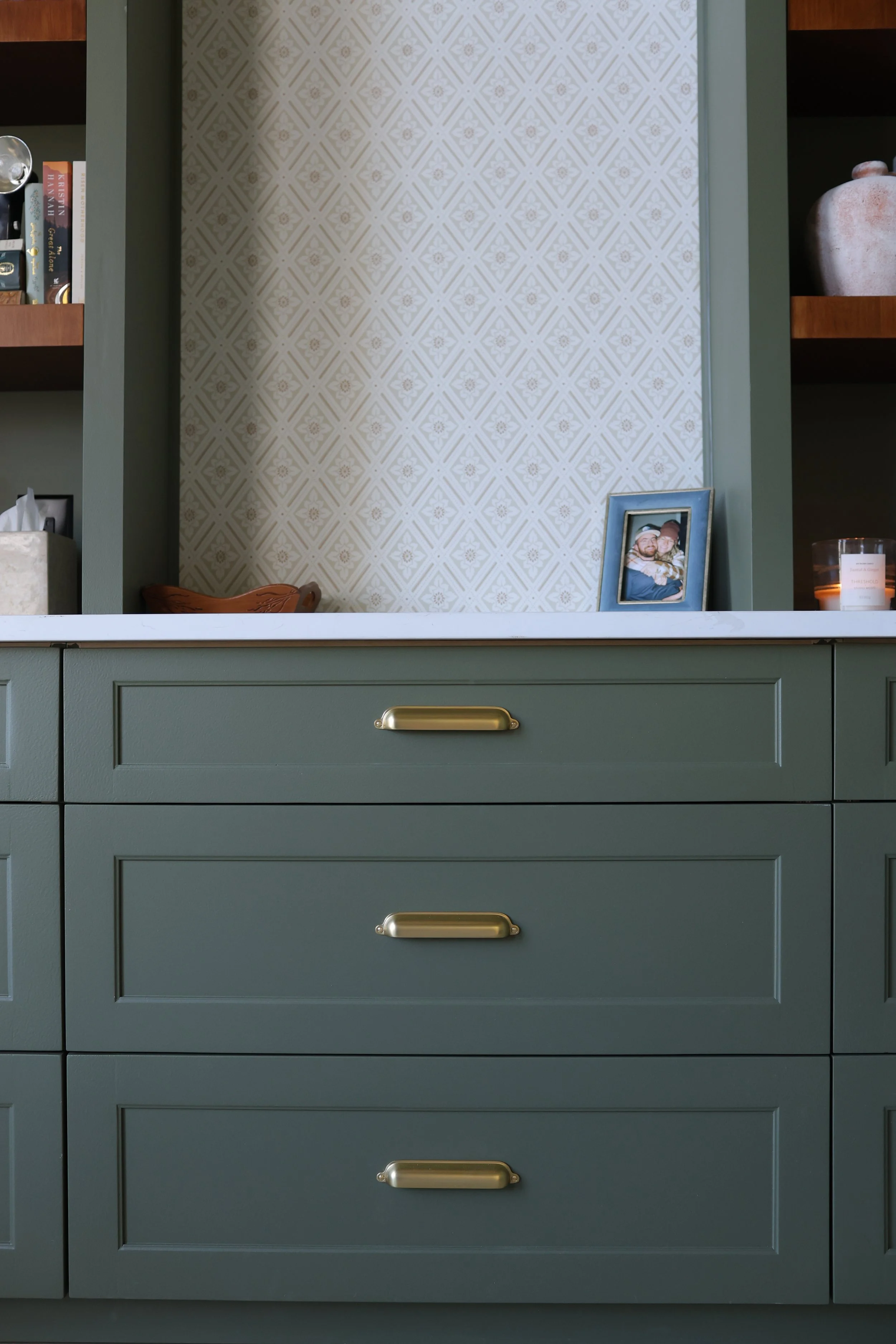 Green kitchen cabinet with three drawers, white marble countertop, framed photo, candles, decorative items, and a patterned wallpaper background.
