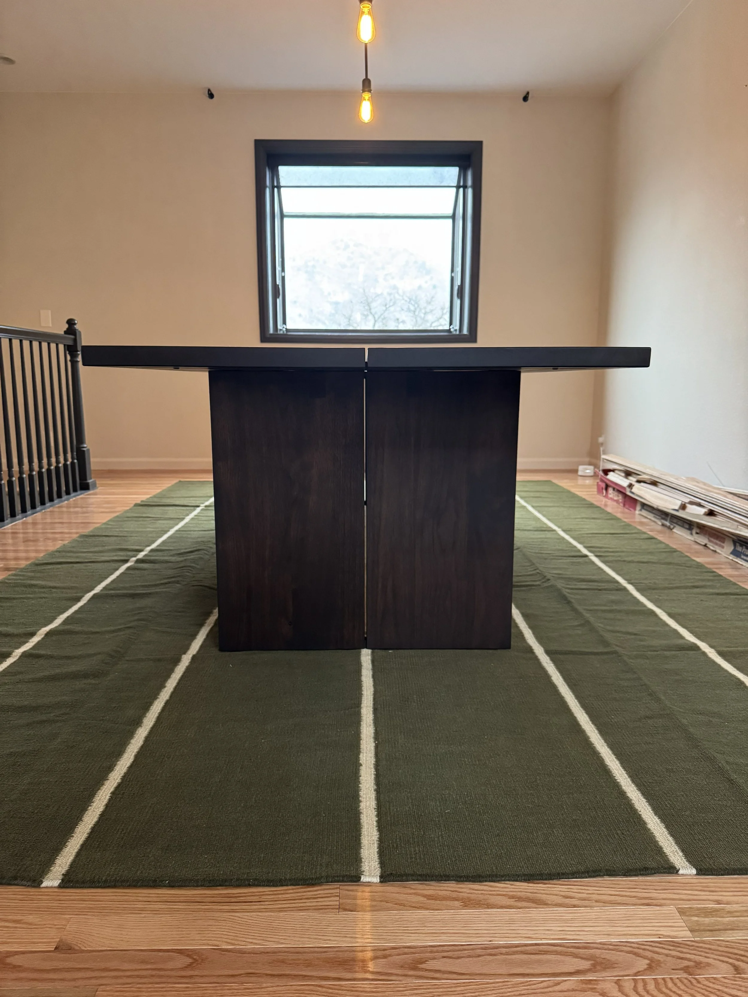 Unfinished room with a dark wooden table on a green striped rug, window in the background, hanging lightbulb fixture, and some construction materials stacked on the right side.