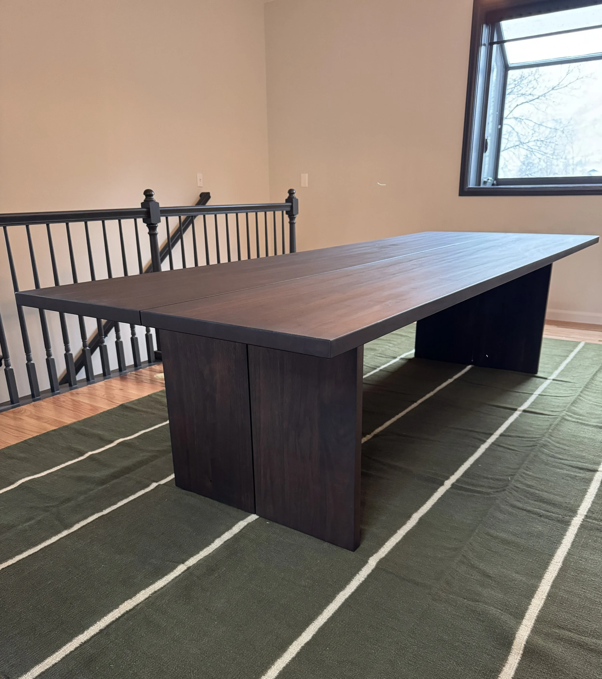A large wooden dining table with a dark finish, placed on a green and white striped rug near a window. There is a stair railing and a blank wall in the background.