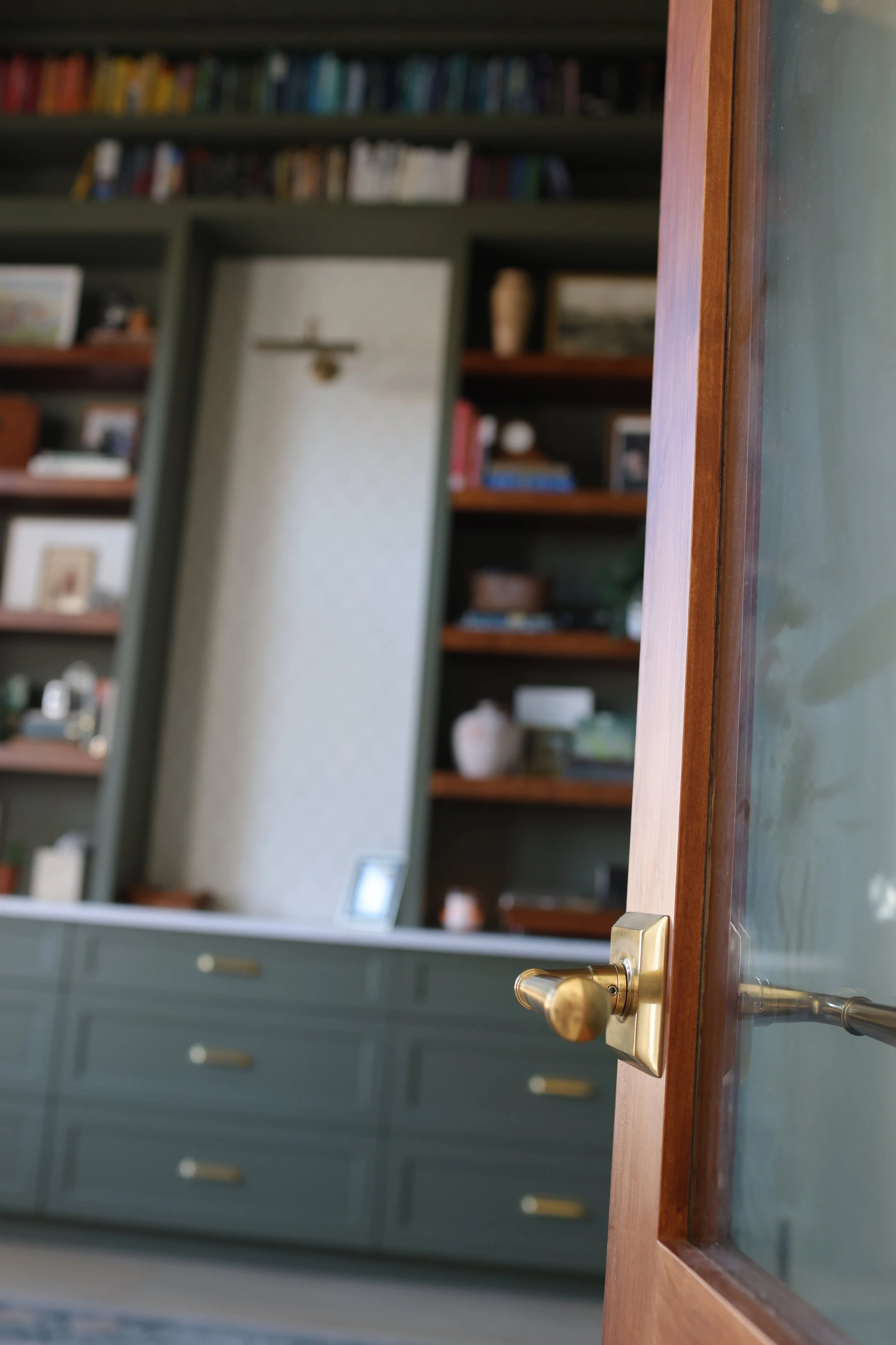 A view through a partially open wooden door with a brass doorknob into a room with dark green built-in bookshelves filled with books and decorative items, and a matching dark green cabinet with gold handles.