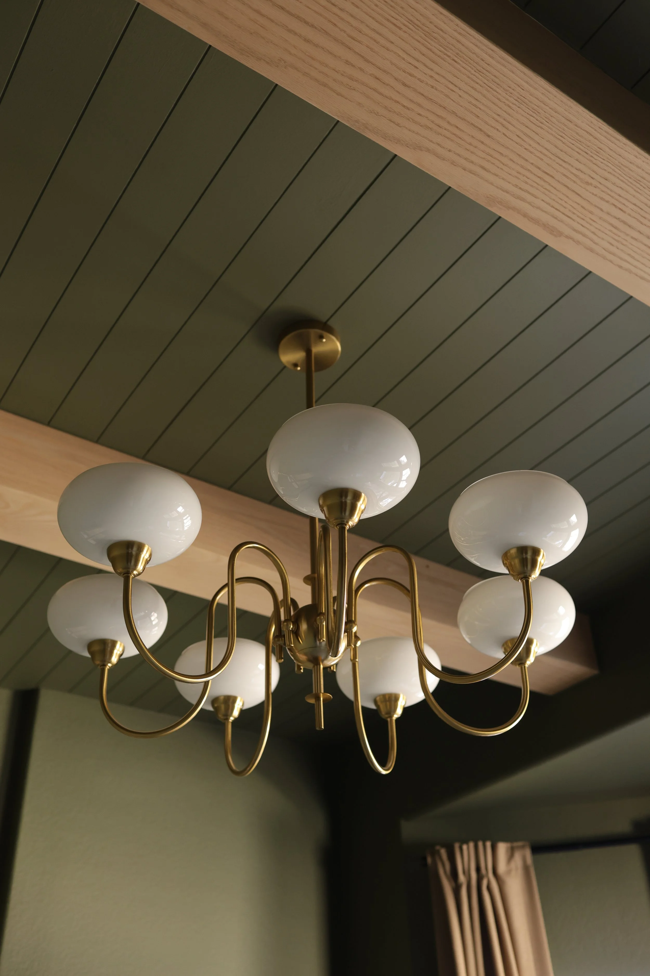 A chandelier with multiple white globe lights and brass arms hanging from a ceiling with green wooden paneling and a wooden beam.