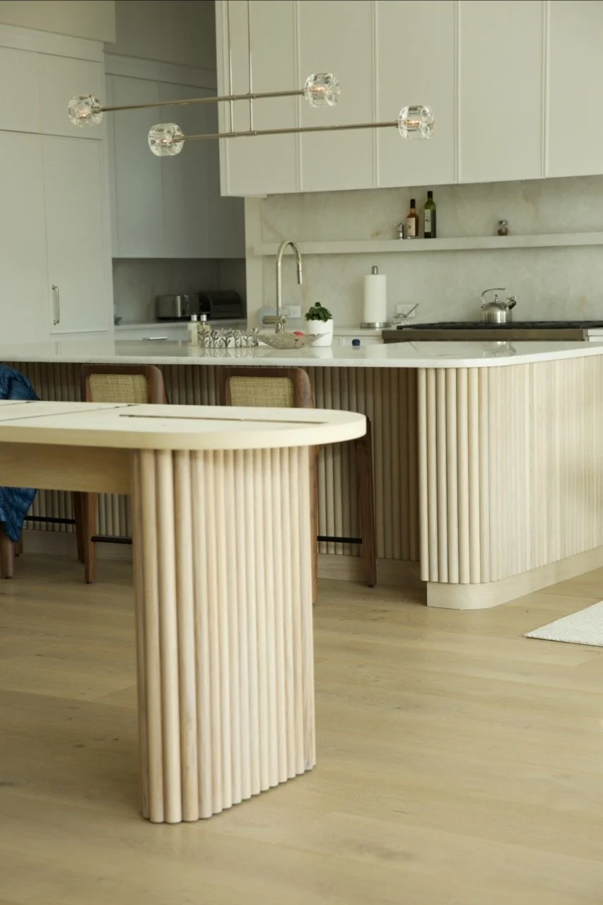 A modern kitchen with white cabinets, a marble backsplash, and a marble countertop. There is a hanging light fixture with glass bulbs, a sink, and some kitchen accessories. Part of a dining table and chairs are visible.