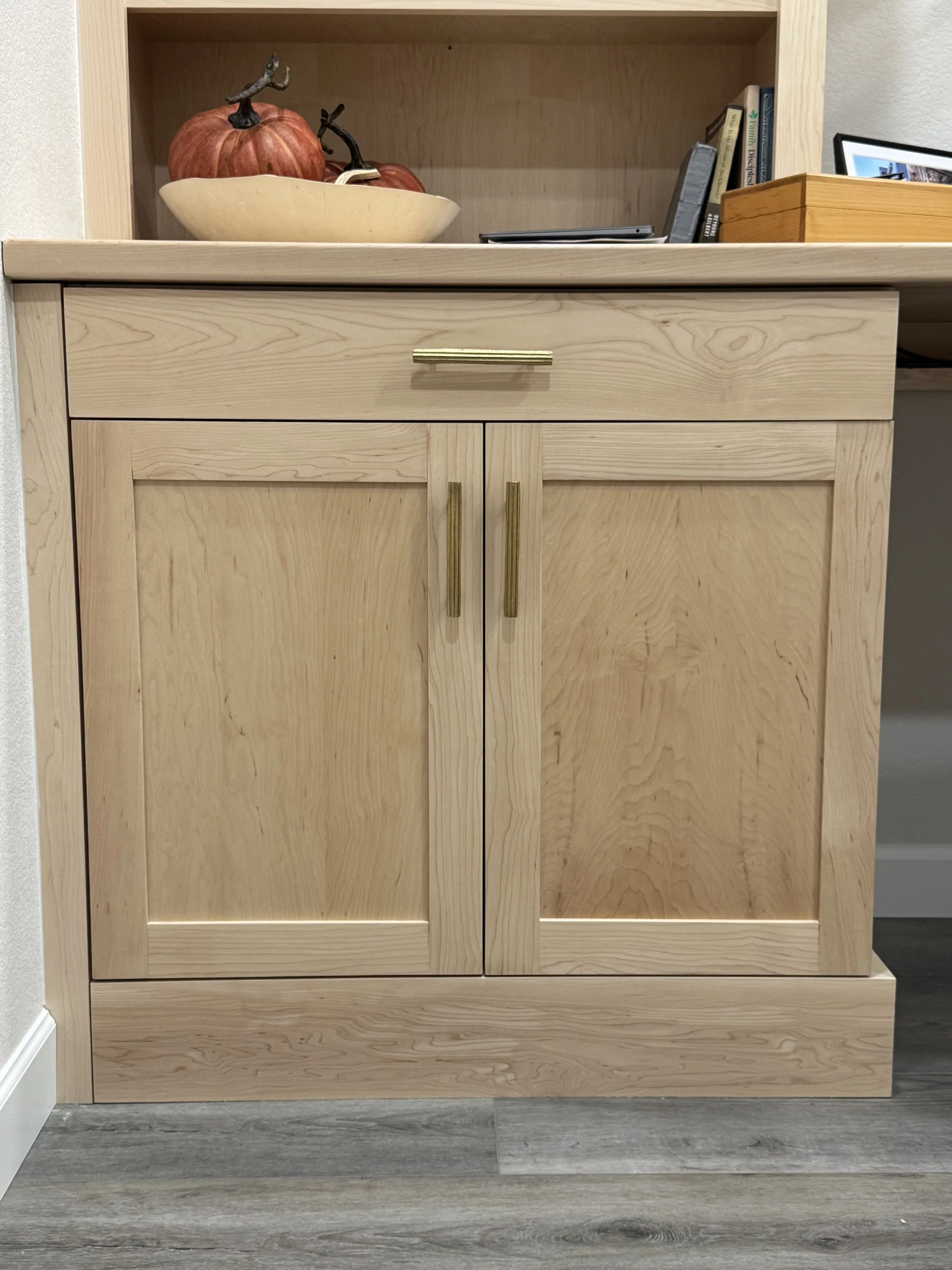 A light wood cabinet with two doors and a single drawer above, set against a wall, with a floor of grayish wood planks.