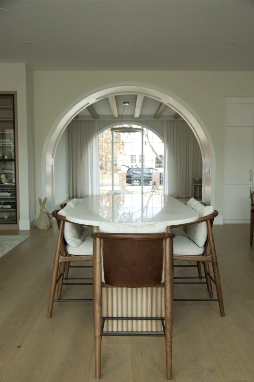 A dining area with a large white marble table surrounded by six chairs, located in front of a sliding glass door leading to the outside, with a view of a driveway and parked cars.