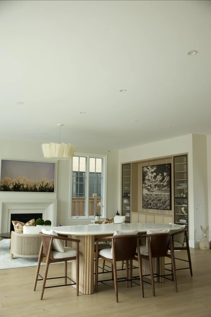 Modern dining and living room with a large round marble table, six chairs, light wood floors, a white fireplace, a framed floral painting, and a window with a wooden fence outside.
