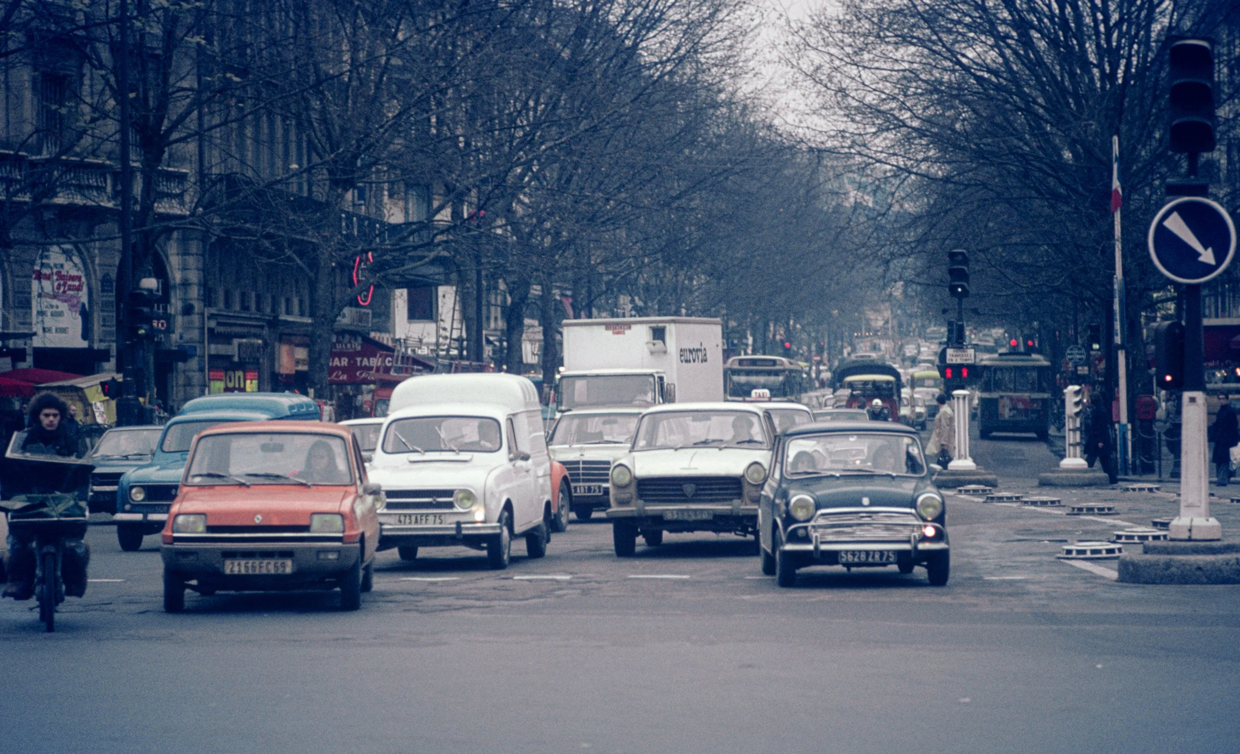 Street scene with vintage cars, a cyclist, trees lining the road, and people on the sidewalks, in an urban area with storefronts and traffic signs.