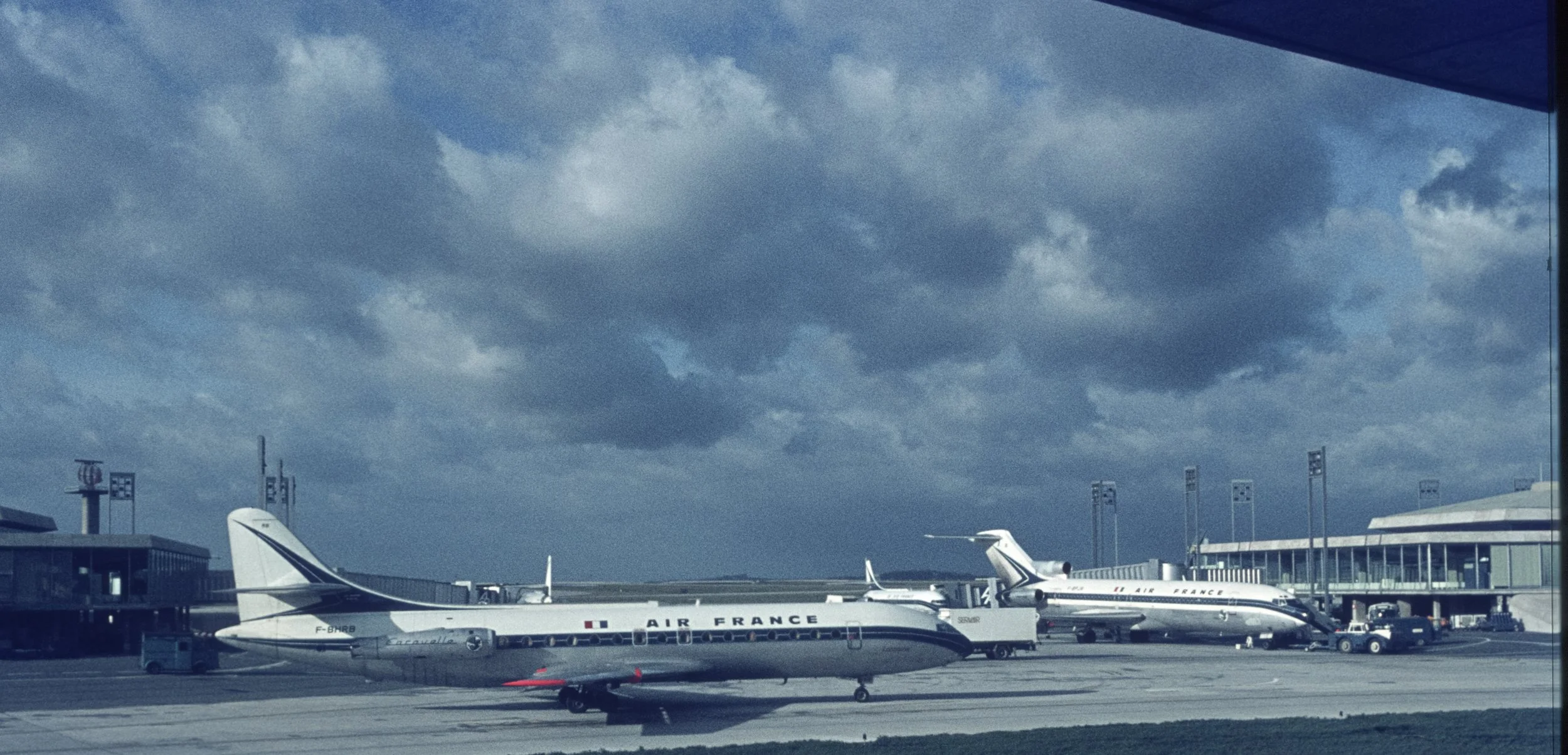 Two Air France airplanes parked at an airport gate under a cloudy sky, with ground vehicles nearby.