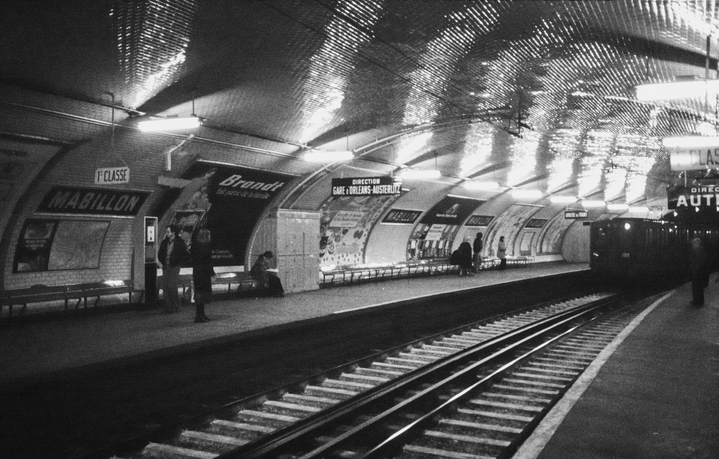 Black and white photo of a Paris Métro station platform with a train arriving, advertisement posters on the wall, and a few passengers waiting.