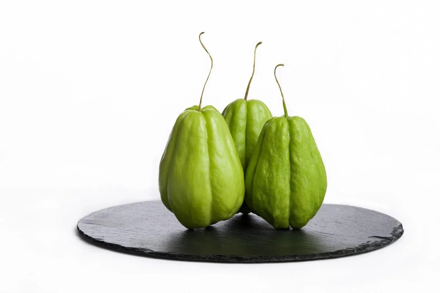 Three chayote squashes with green skin and long stems on a black slate serving platter against a white background.