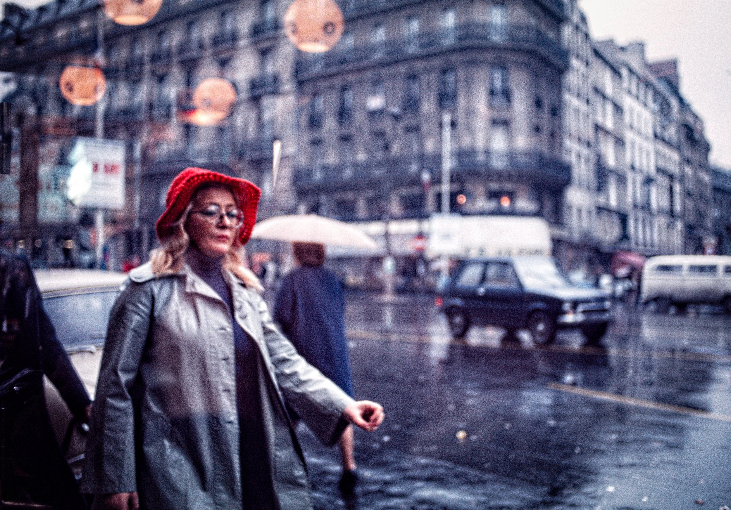 A woman wearing glasses, a red knit hat, a gray coat, and a purple shirt walking on a rainy city street holding an umbrella in the background.