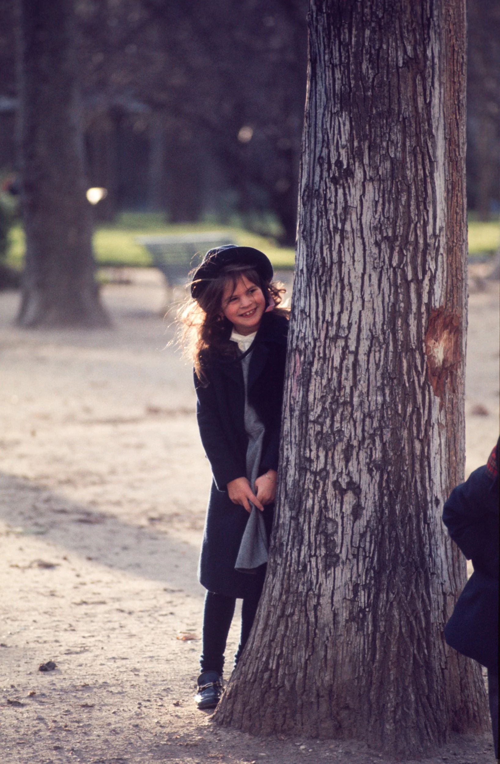 A young girl with dark hair, wearing a dark coat and hat, peeks out from behind a large tree and smiles.