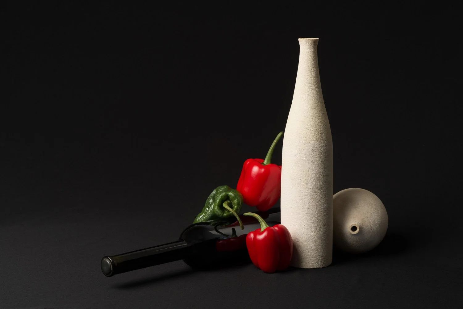 Still life with red and green peppers, a white textured bottle, a round ceramic vessel, and a black object on a dark surface.