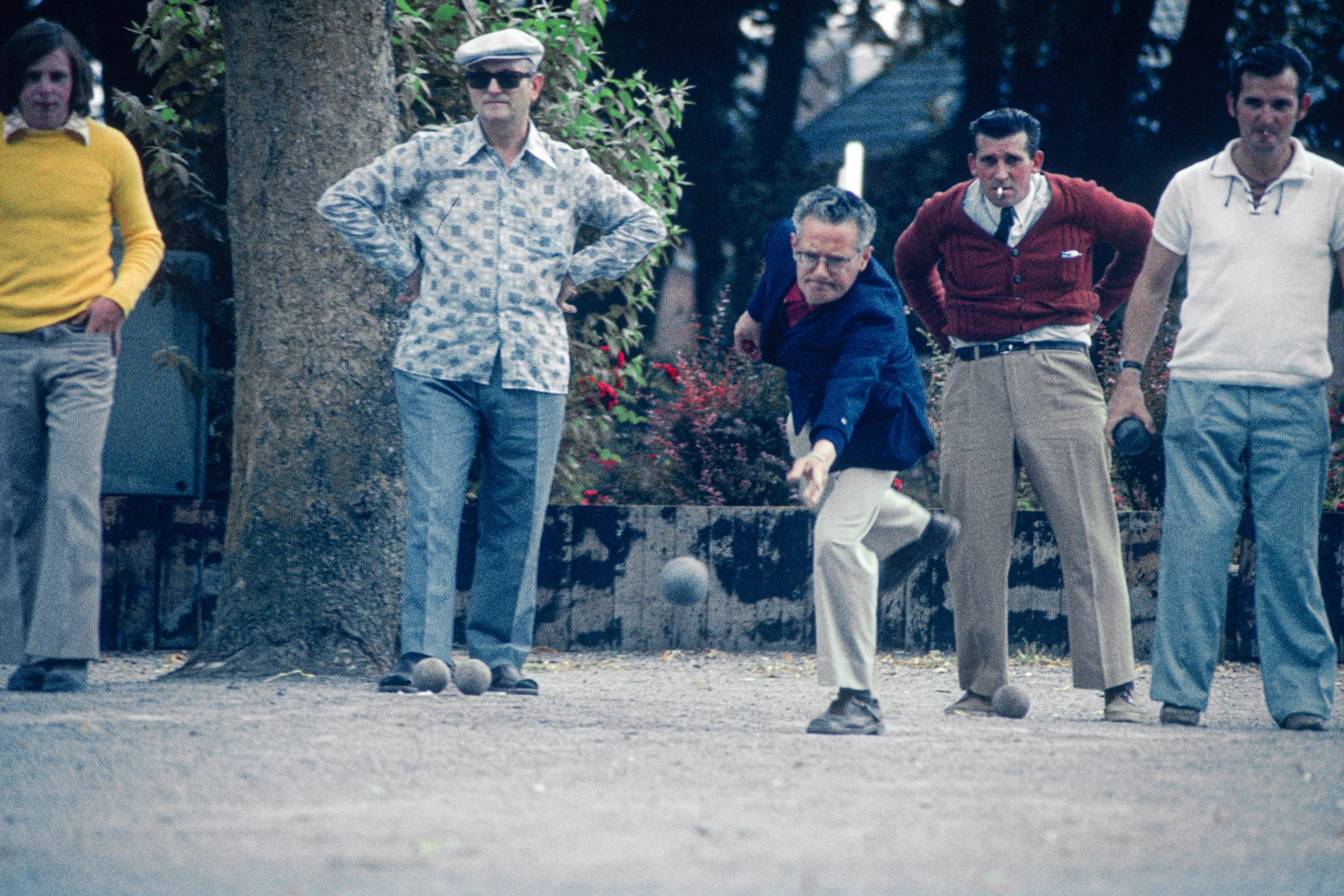 Group of five men standing outdoors, one in mid-throw of a bocce ball, with three others watching and one partly visible on the far left.