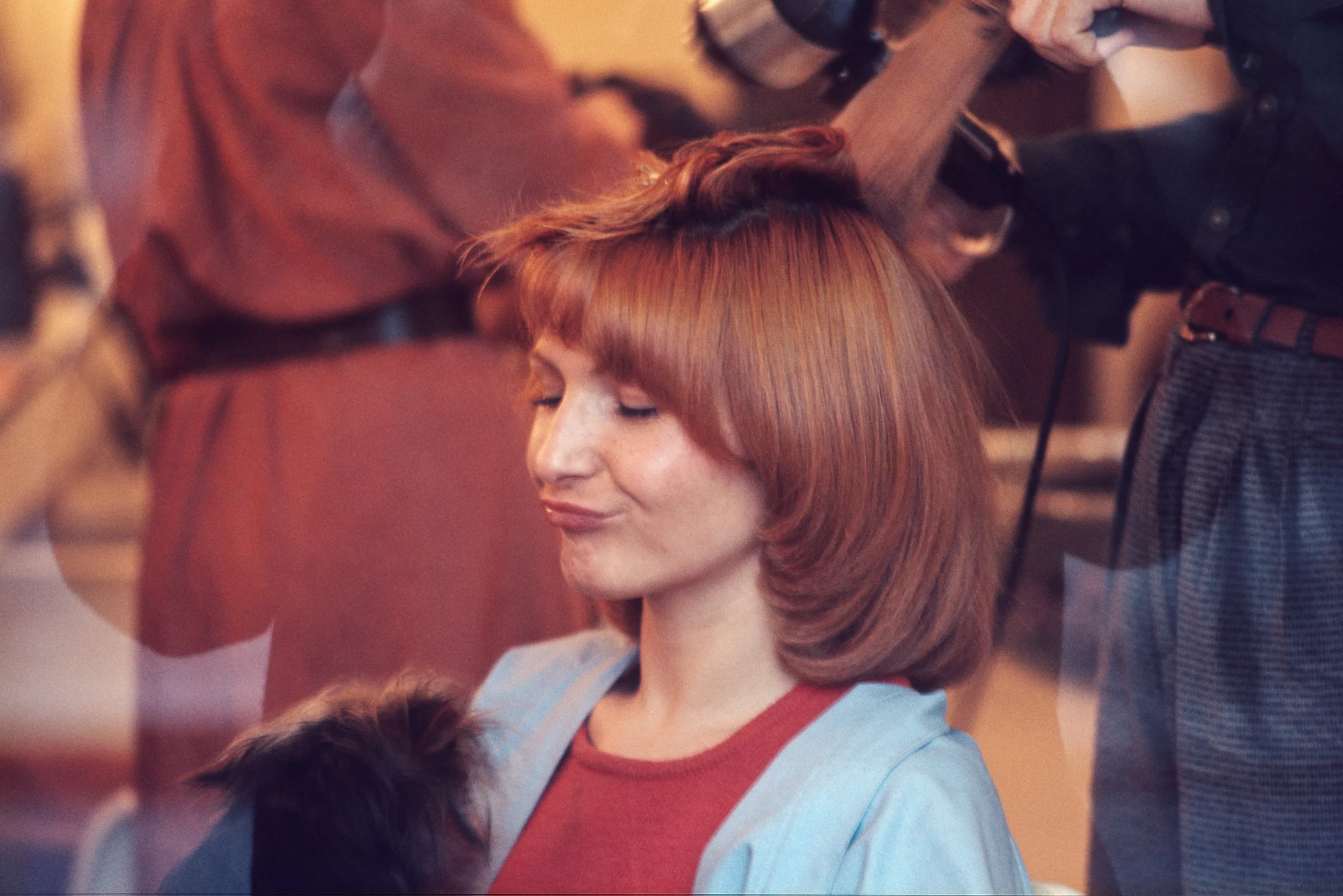 A woman  with red hair and a light blue blazer and a little dog sitting with her eyes closed while a hairstylist trims her hair.