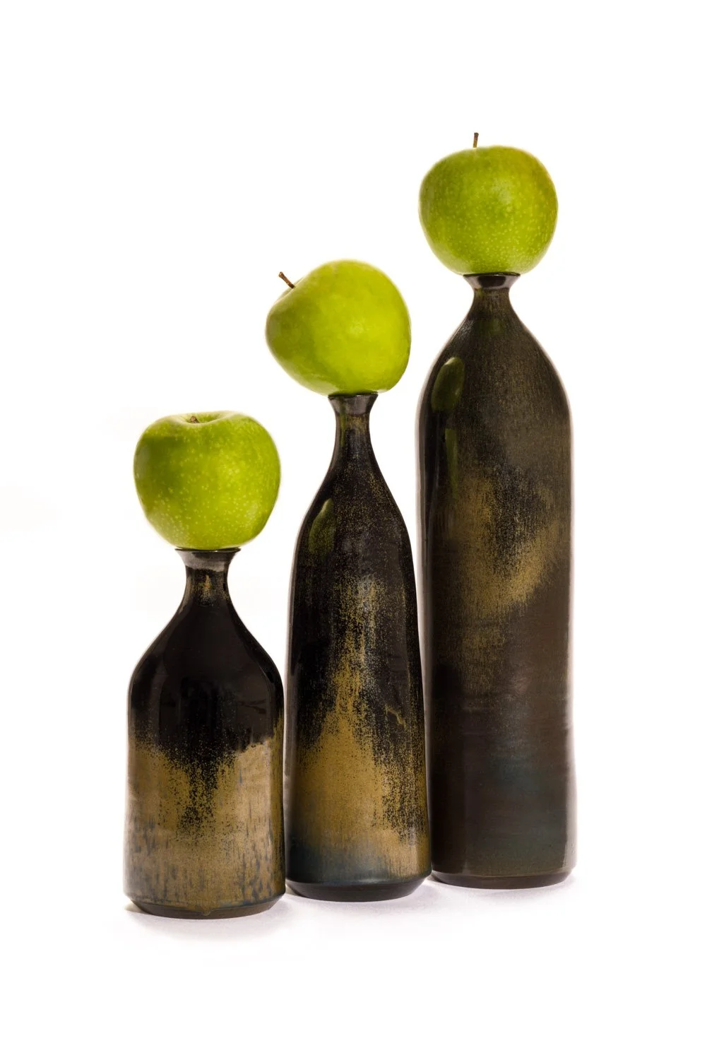 Three black ceramic vases with a distressed finish, each topped with a green apple, arranged in increasing height from left to right.