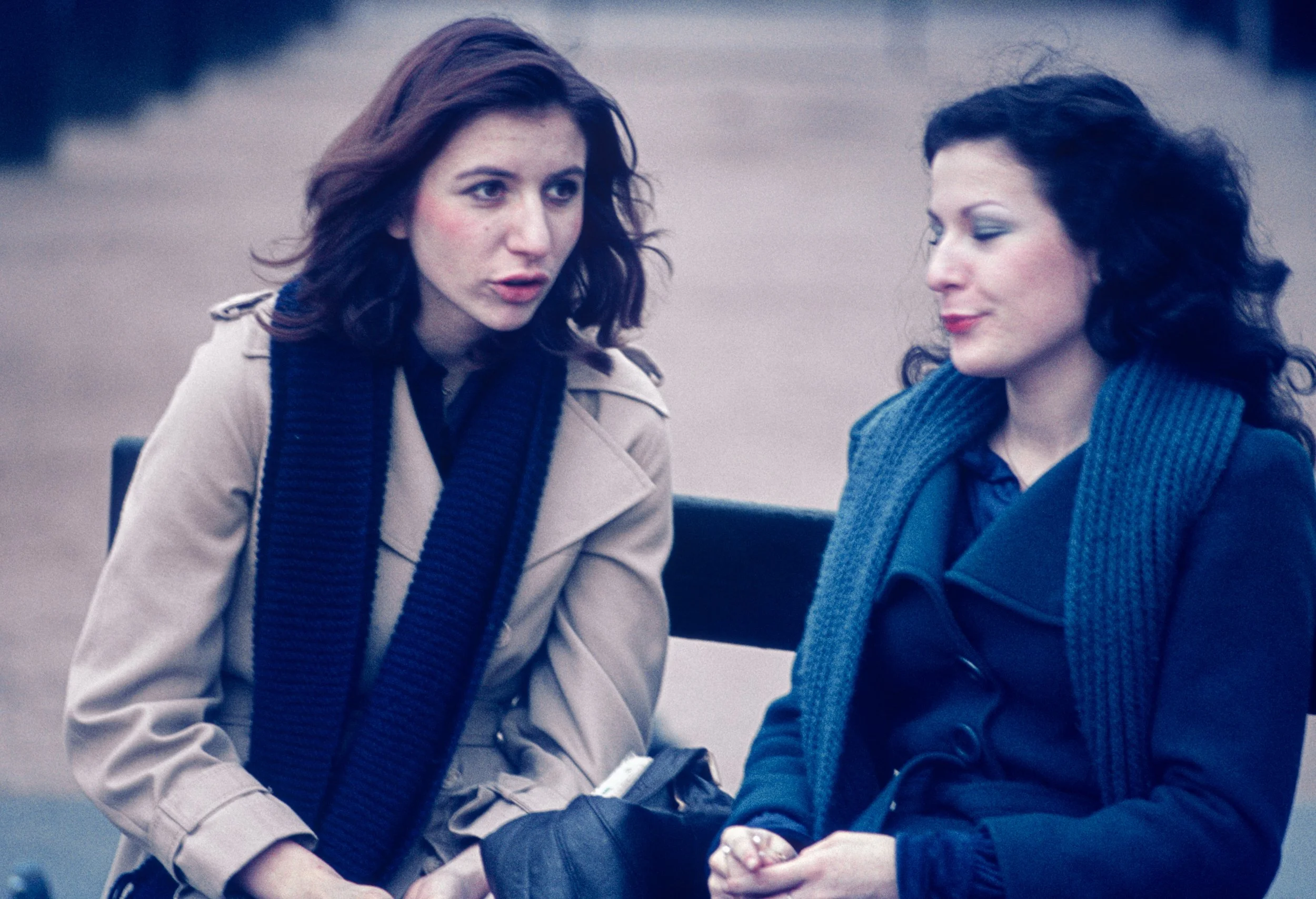 Two women sitting on a park bench engaged in conversation, one with brown hair and the other with black hair, both wearing coats and scarves.