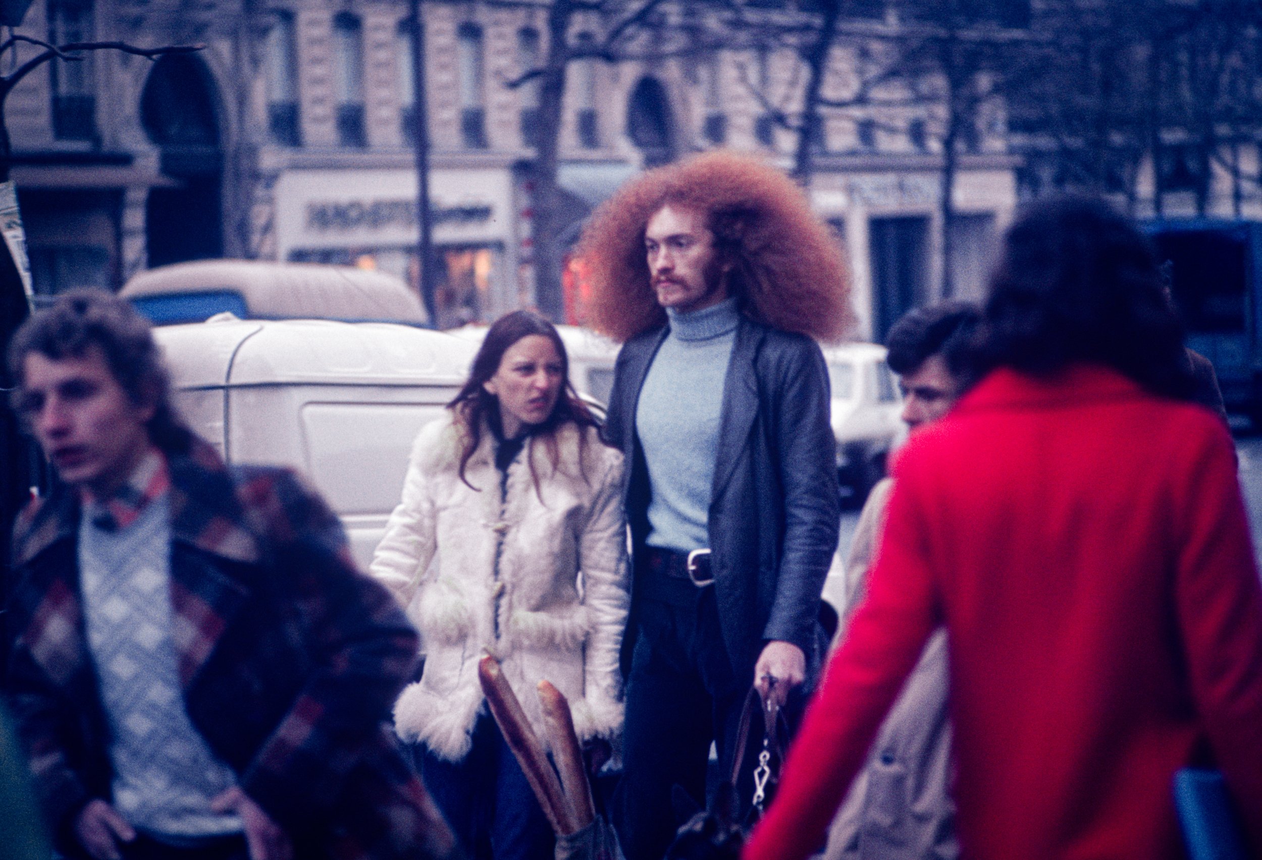 A street scene with several pedestrians, including a tall man with long, curly red hair wearing a gray turtleneck and black leather jacket, walking among other people in colorful coats on an urban street with cars and buildings in the background.