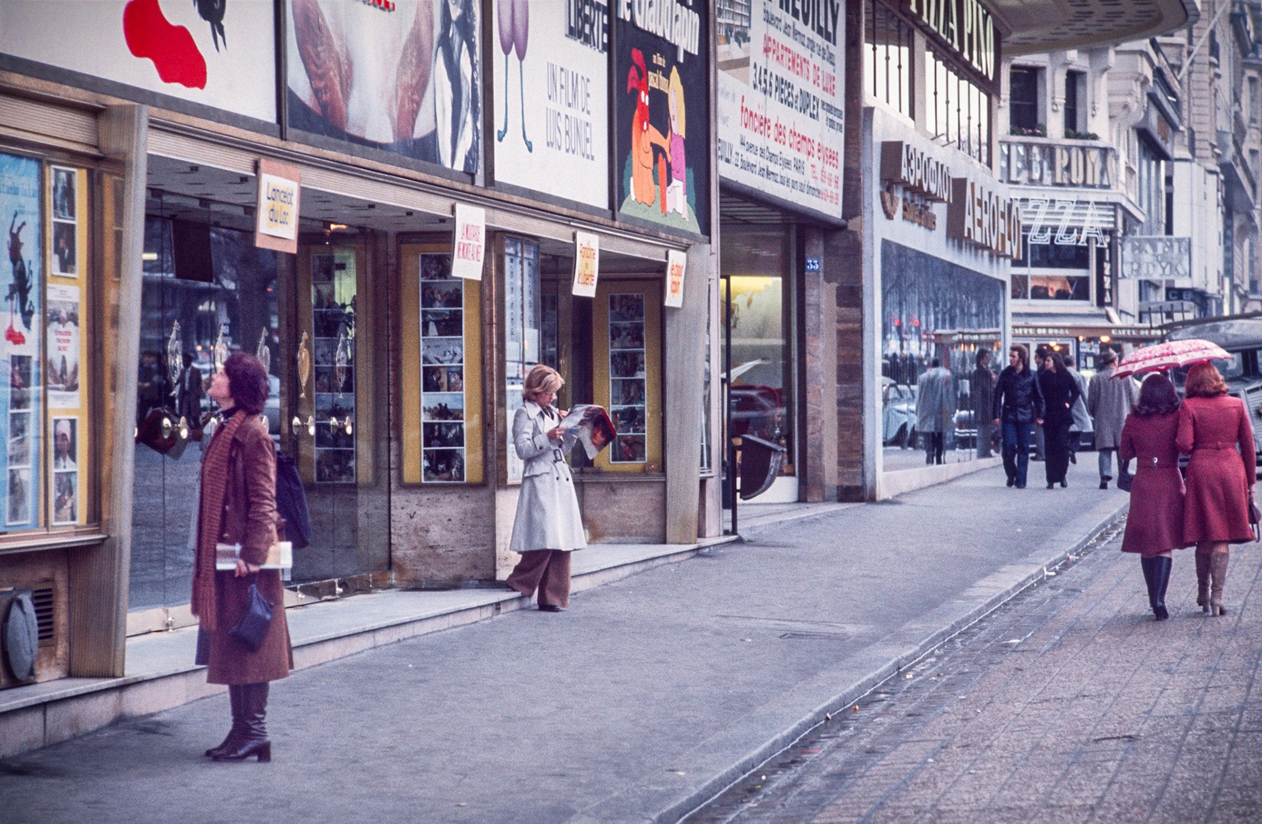 People walking on a city sidewalk in front of stores and cinema with large signs and colorful posters, some holding umbrellas, during cloudy weather.