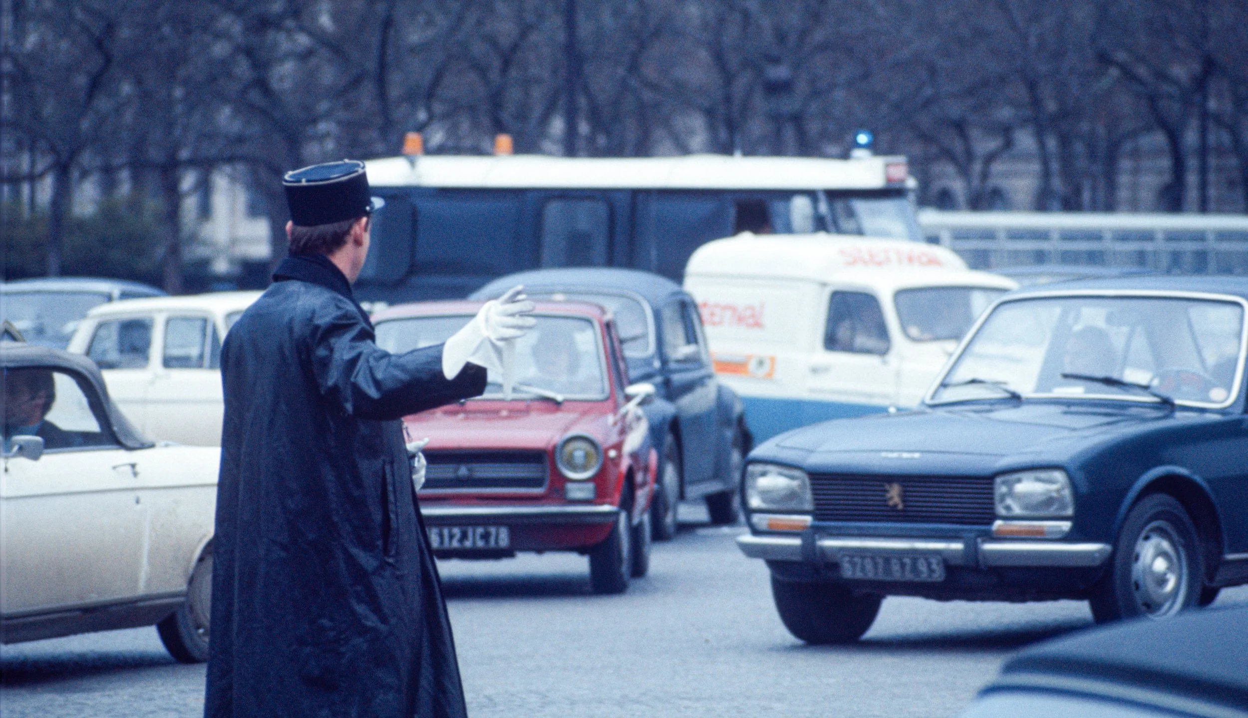 A traffic police officer in a black hat and coat, with white gloves, stands on a street directing cars. Several vintage cars and an ambulance are visible in the background.