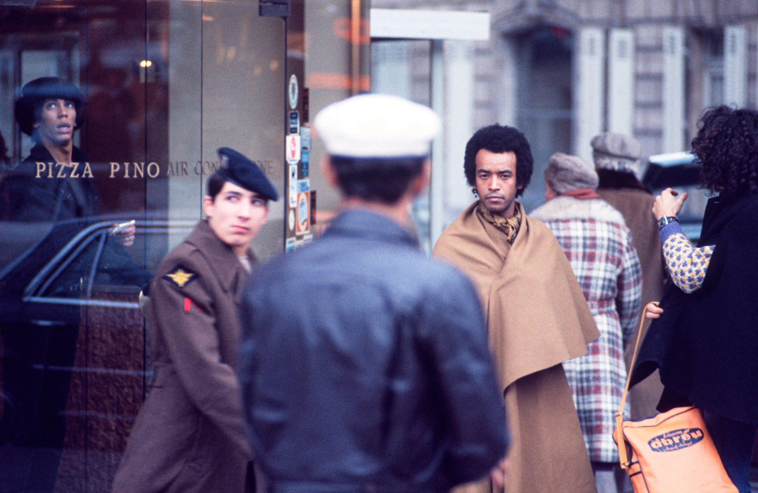 A group of people standing outside a storefront with a sign that reads 'Pizza Pino.' A woman is inside the shop looking out through the glass, and several women and men are gathered on the sidewalk, some wearing period clothing including berets and c