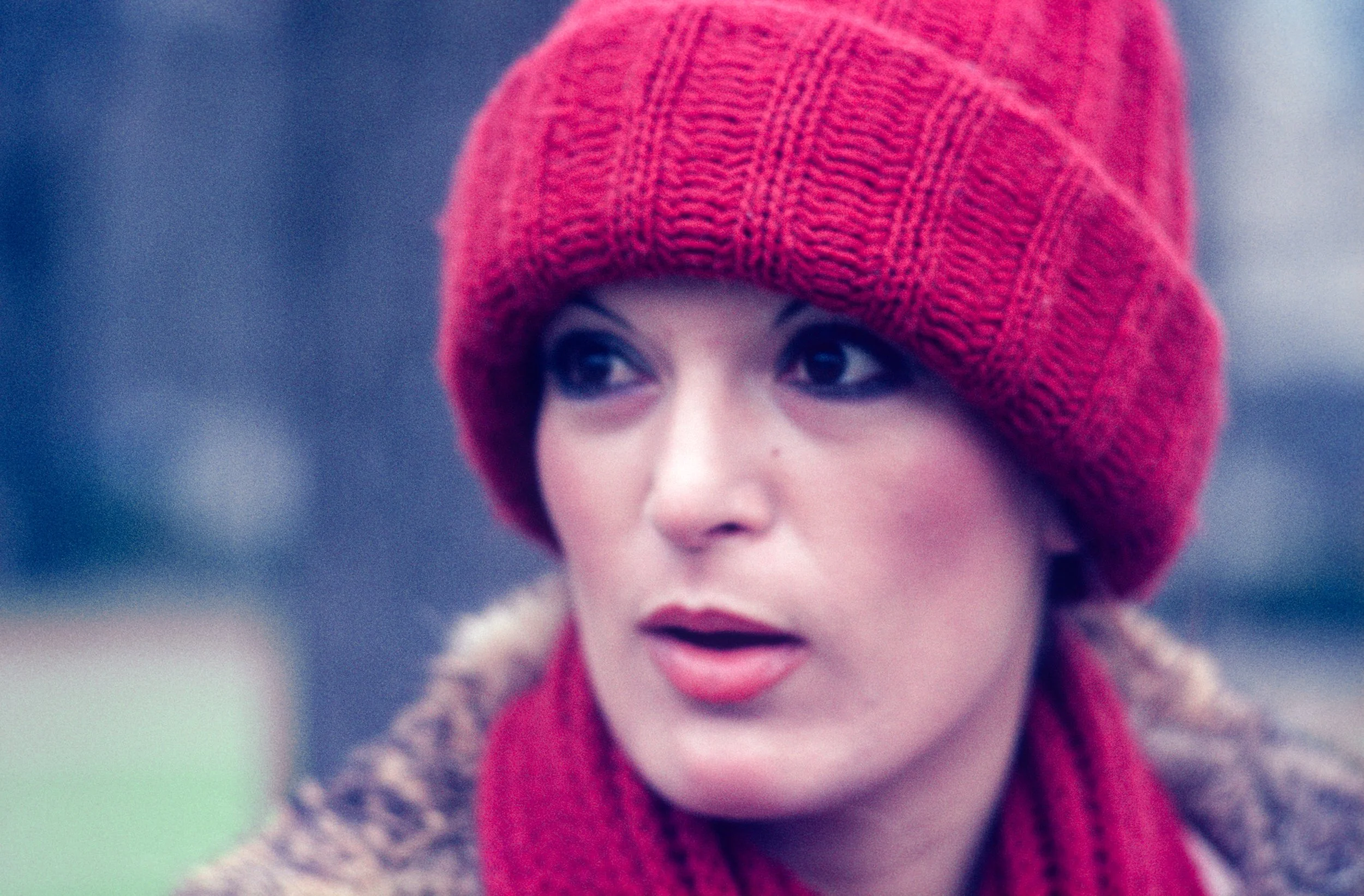 A woman wearing a red knit hat and scarf, with a brown coat, outdoors with a blurred background.