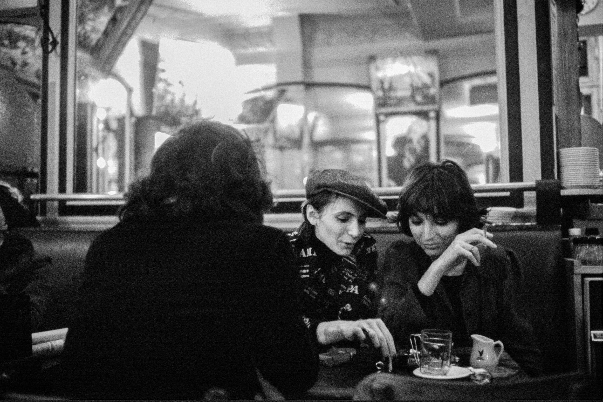 Two women and a man sitting at a table in a restaurant or café, engaged in conversation. The photo is in black and white, with a window or mirror behind them reflecting the interior.