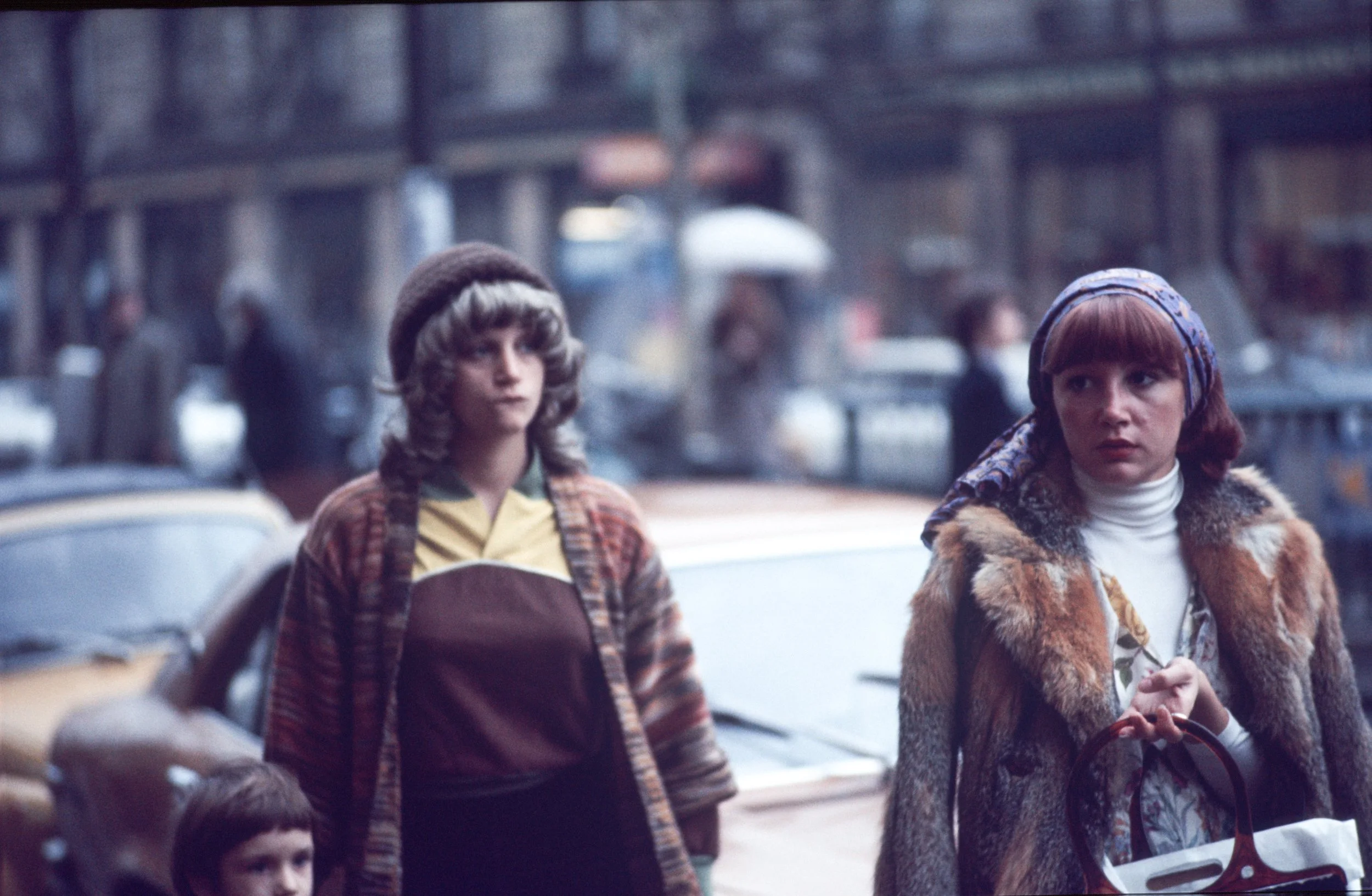 A woman with red hair wearing a headscarf and fur coat stands on a city street, holding a shopping bag, with two children nearby. The background shows a busy urban scene with cars and pedestrians.