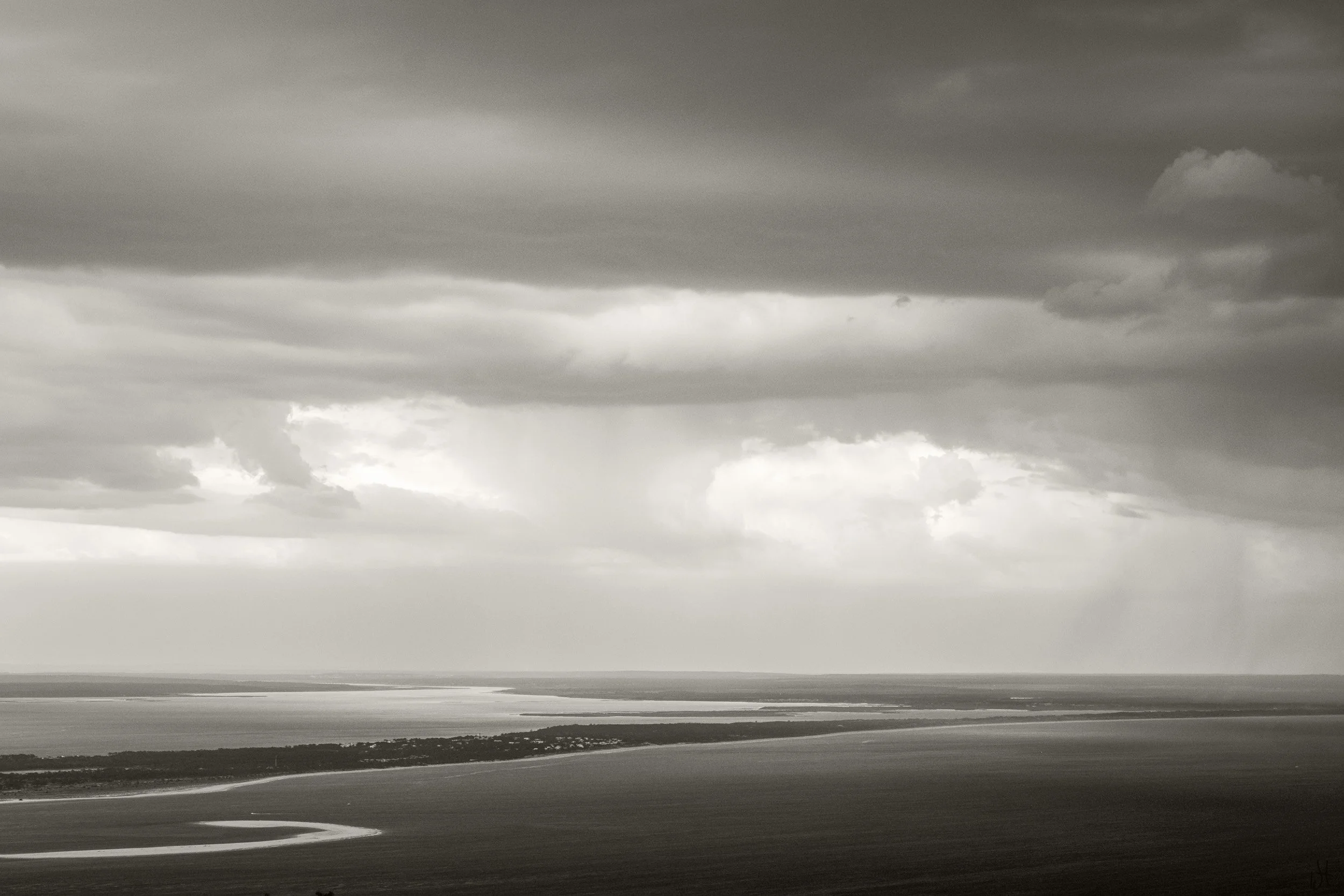 A black and white landscape photograph showing a wide river or lake with a small winding waterway in the foreground, a strip of land with trees in the middle ground, and a cloudy sky with dark storm clouds overhead.