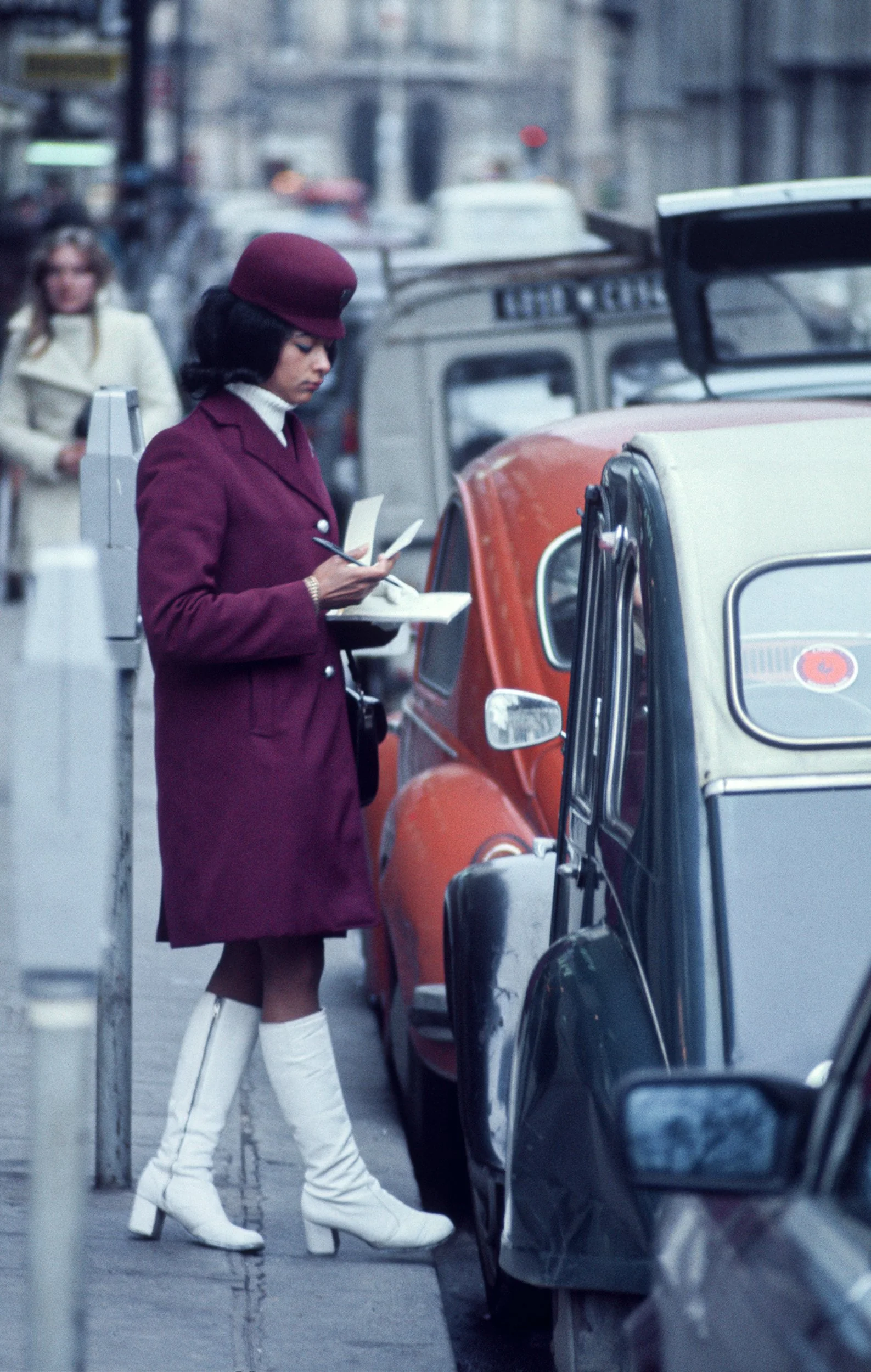 A woman in a burgundy coat, matching hat, white turtleneck, and white knee-high boots is writing on a notepad while standing on a city sidewalk next to parked cars.
