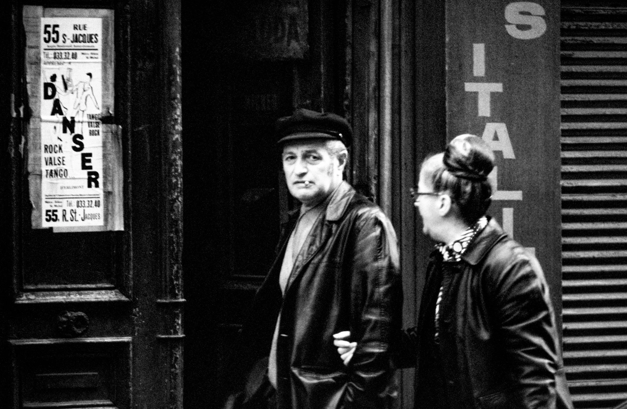 A black-and-white photo of a man and woman standing outside a dance studio. The man is wearing a leather jacket and a cap, with a cigarette in his mouth, looking directly at the camera. The woman is wearing glasses and a leather jacket, turned slight