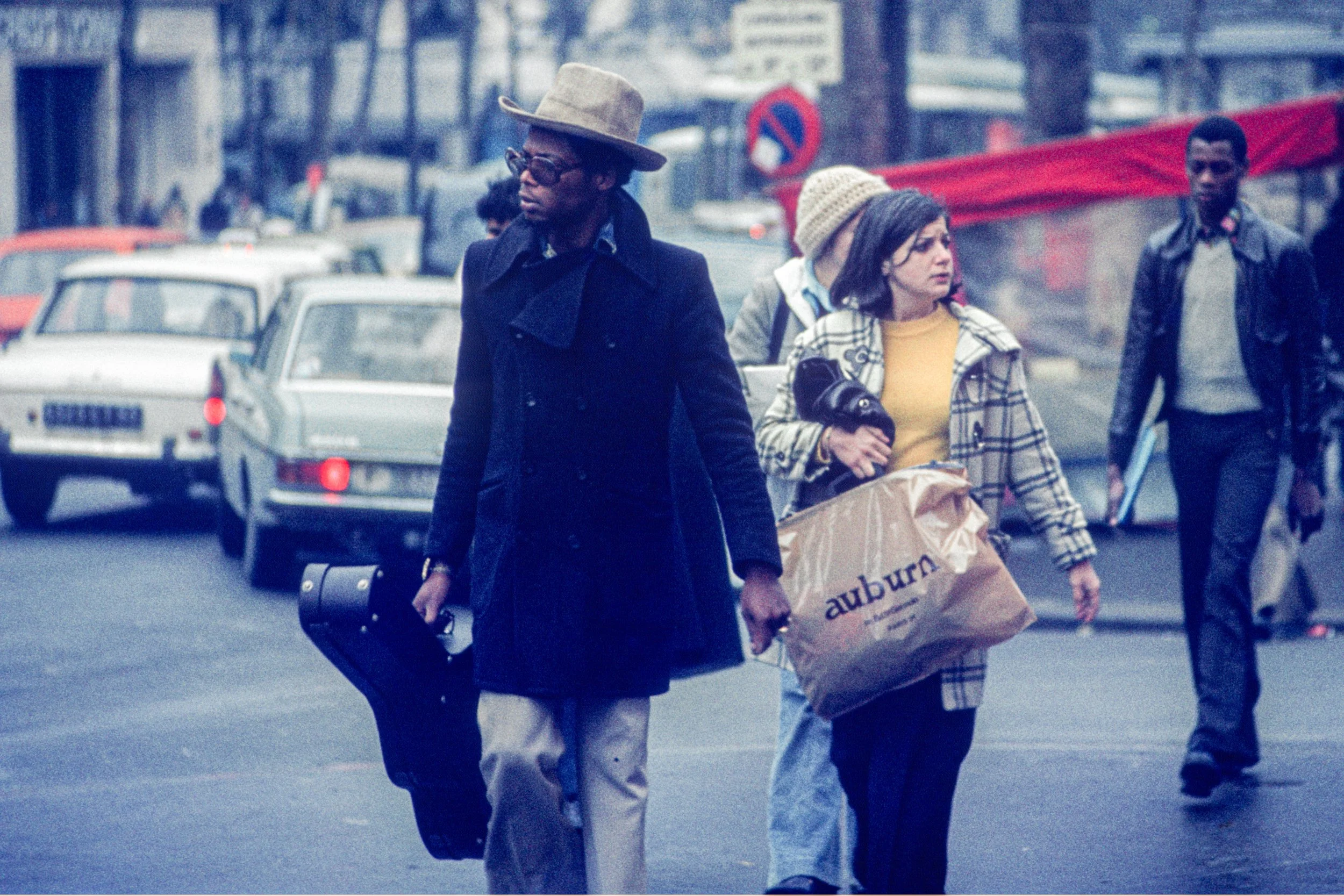 A man and a woman walk across a street, with the man carrying a guitar case, and the woman holding a paper shopping bag. Other pedestrians and parked cars are visible in the background.