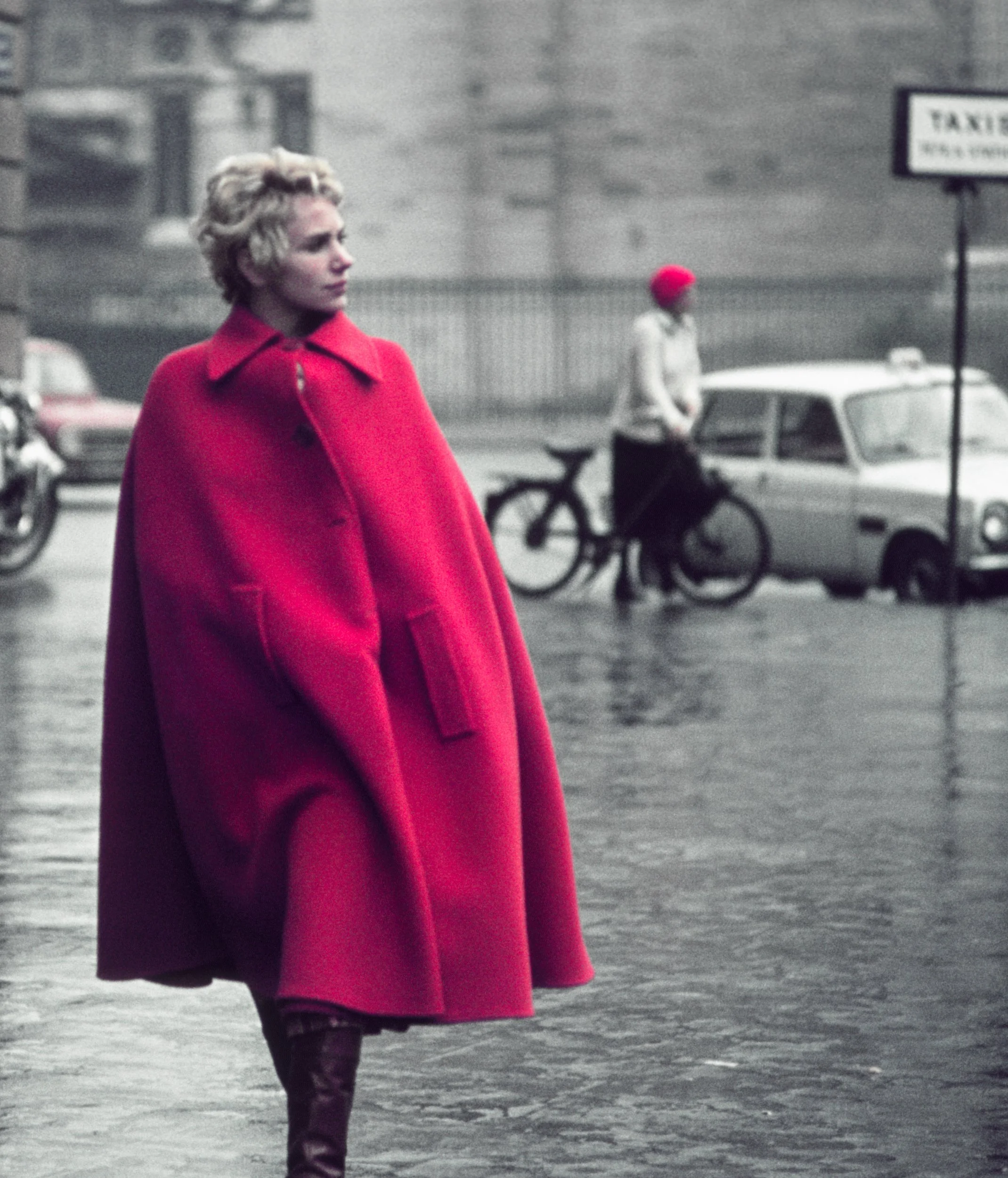 A woman in a bright red coat standing on a street with a blurred background that includes a person riding a bicycle, a parked car, and a sign that reads 'TAXIS'. The woman has short blonde hair and is gazing to her left.