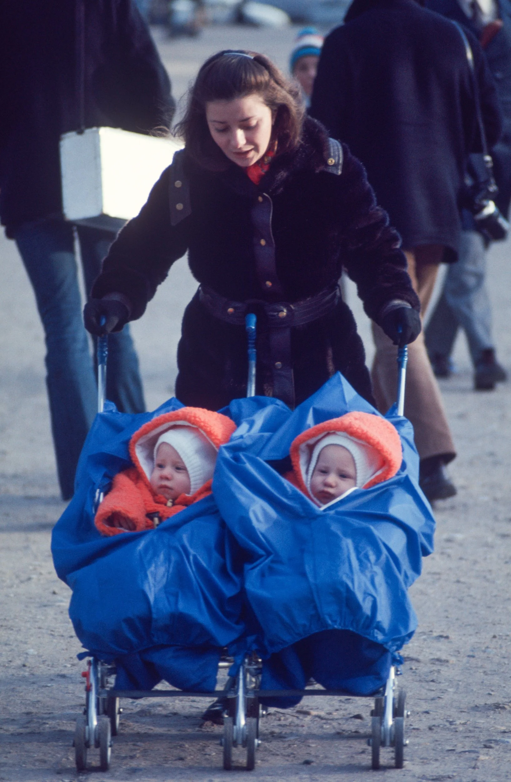 A woman is pushing a double stroller with two babies wearing warm clothing, including orange hats and blankets, in an outdoor setting with other people in the background.