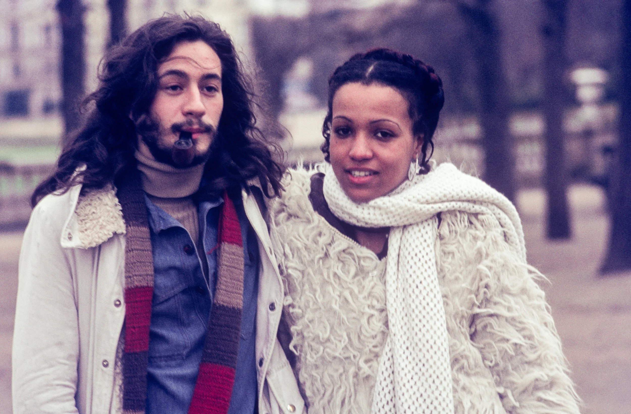 A man with long, curly hair, a beard, and a pipe, standing next to a woman with dark curly hair, both outdoors in a park.