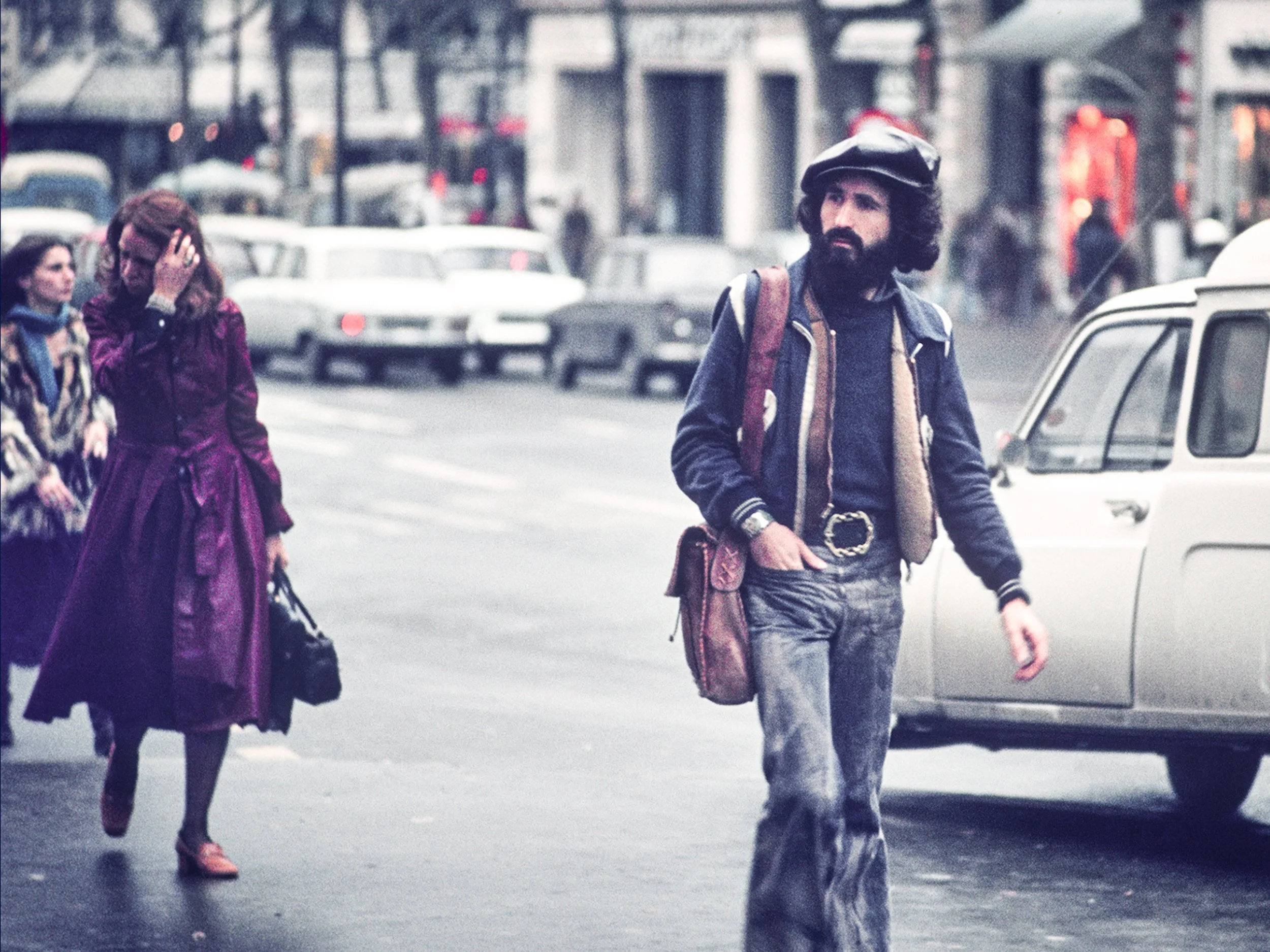 A man with a beard, wearing a hat, jacket, and jeans, walks on a city street next to a parked car, while women in colorful coats walk in the background.