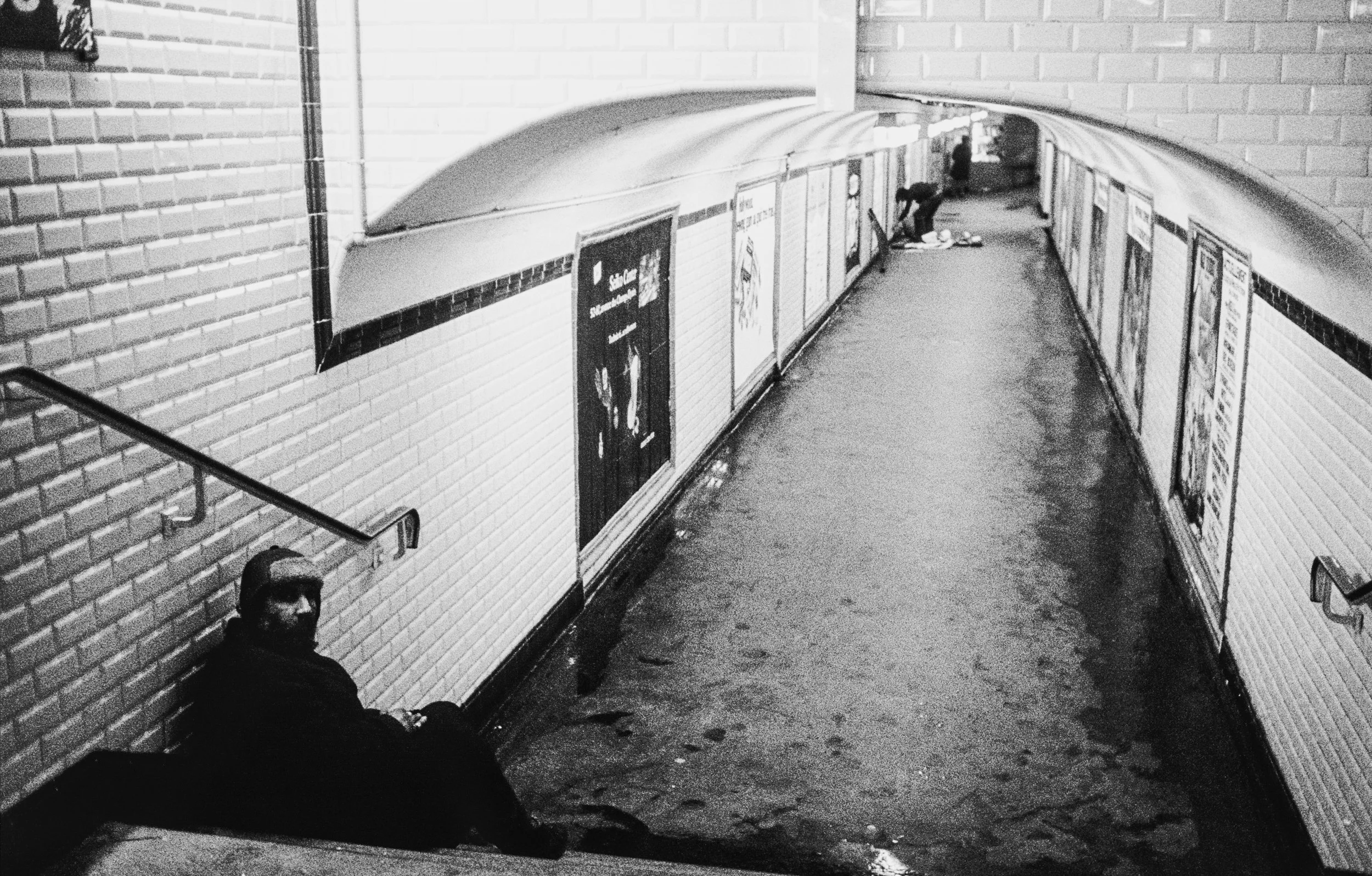 An underground subway tunnel with two men, one sitting against a brick wall and another further down near the wall, with advertisements on the tunnel walls.