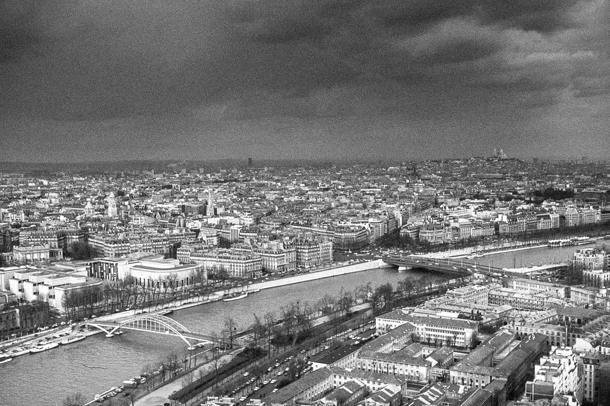 A black and white aerial view of Paris, France, showing the Seine River with bridges, surrounding buildings, and overcast cloudy skies.