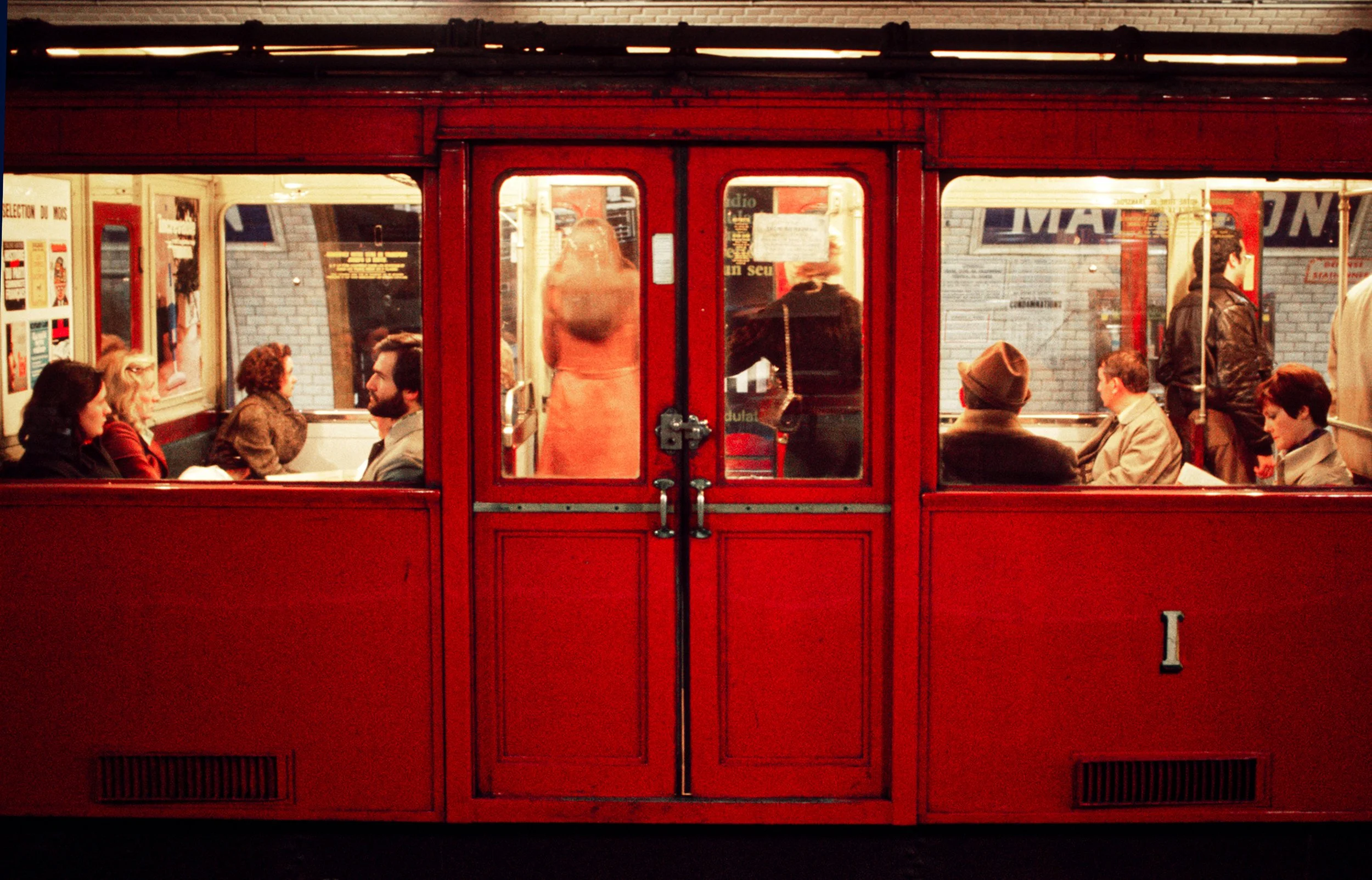 People sitting in a vintage red tram, viewed through closed door, with outdoor city scene in background.