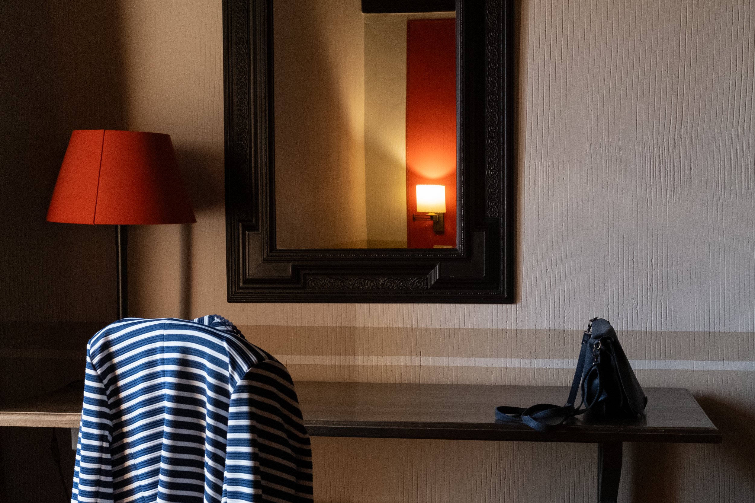 A quiet still life captured in a hotel room: a small table sits beneath a mounted mirror, bathed in the warm glow of a red lampshade. A handbag rests casually on the table, hinting at a moment of pause between comings and goings. The composition evok