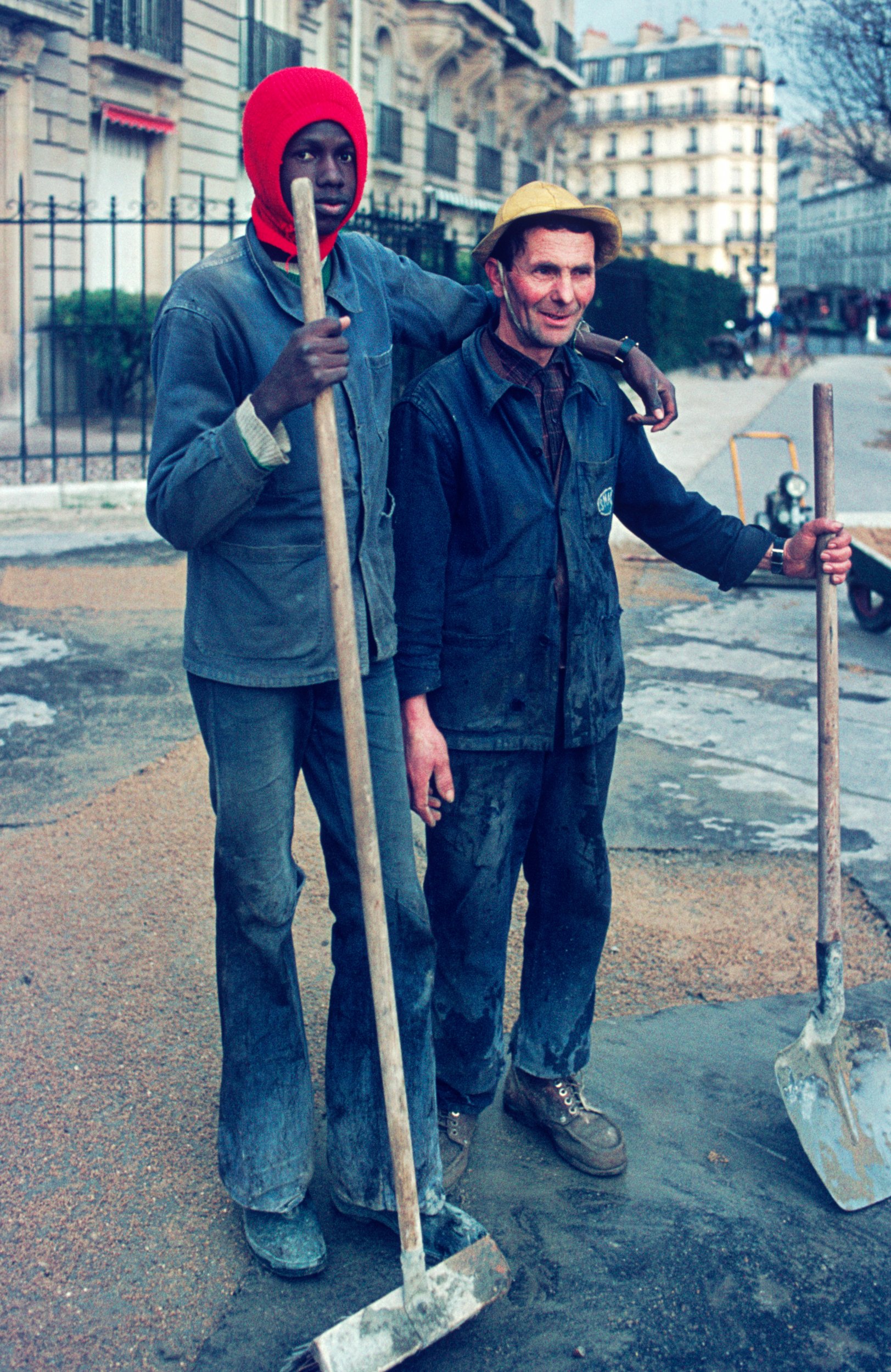 Two construction workers, one tall and dark-skinned wearing a red beanie, and the other shorter and light-skinned with a yellow hat, standing on a street with buildings in the background. They are holding shovels, and the ground around them appears t