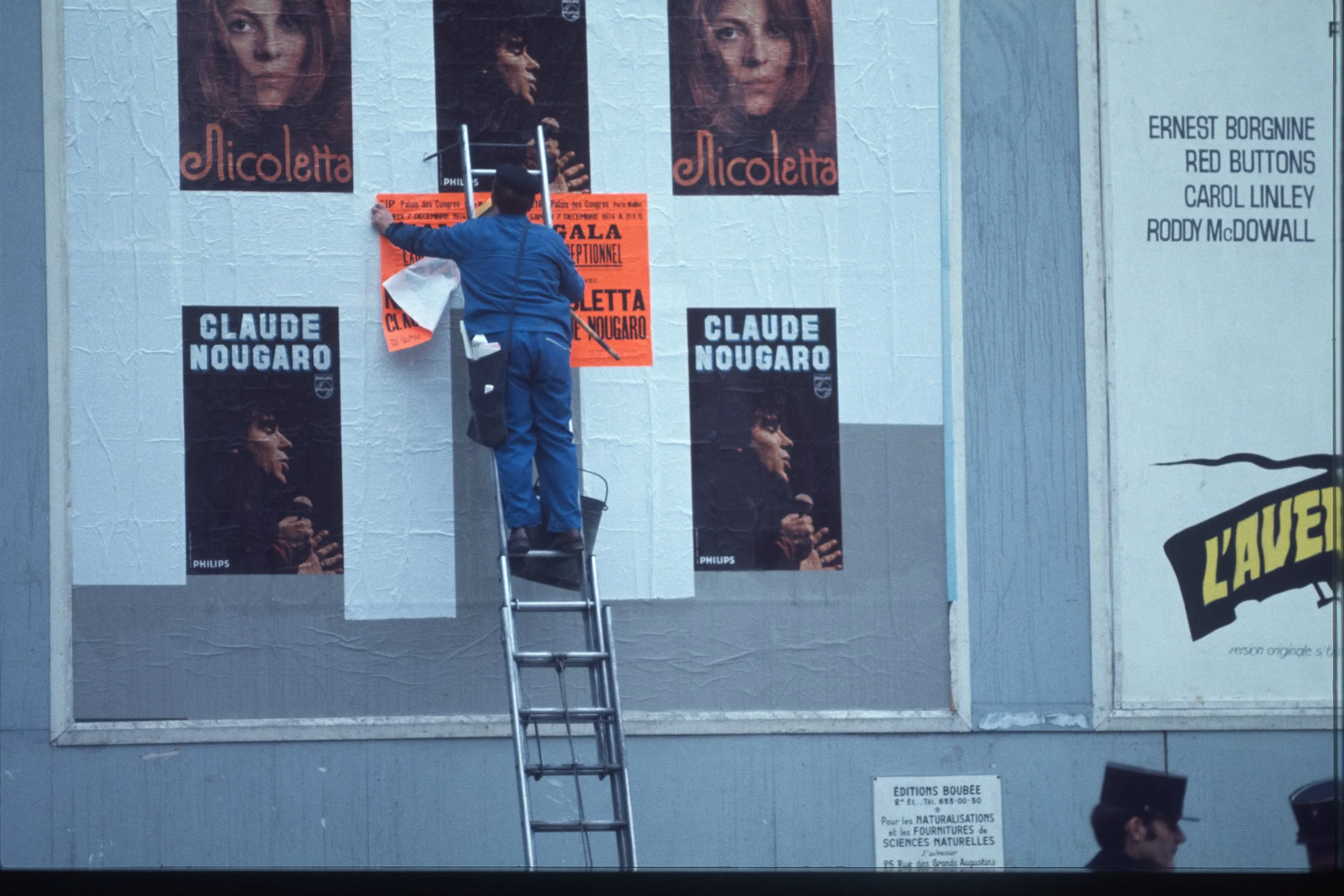 A person on a ladder attaching a poster to a wall with other posters and advertisements. The wall displays posters of a woman named Nicolette and credits to Ernest Borgnine, Red Buttons, Carol Linley, and Roddy McDowall.