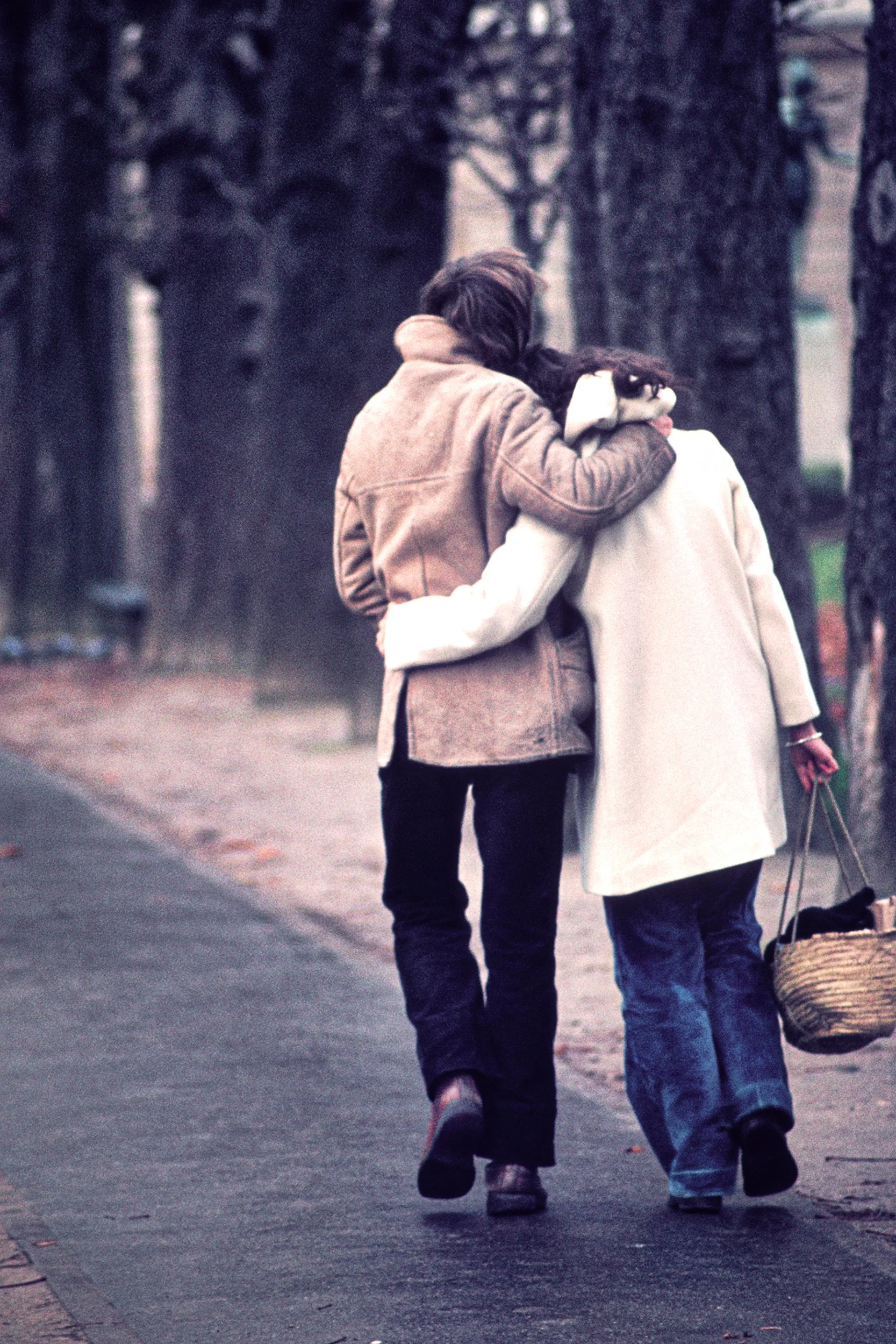 Two women walking arm-in-arm down a tree-lined sidewalk, with one holding a basket.