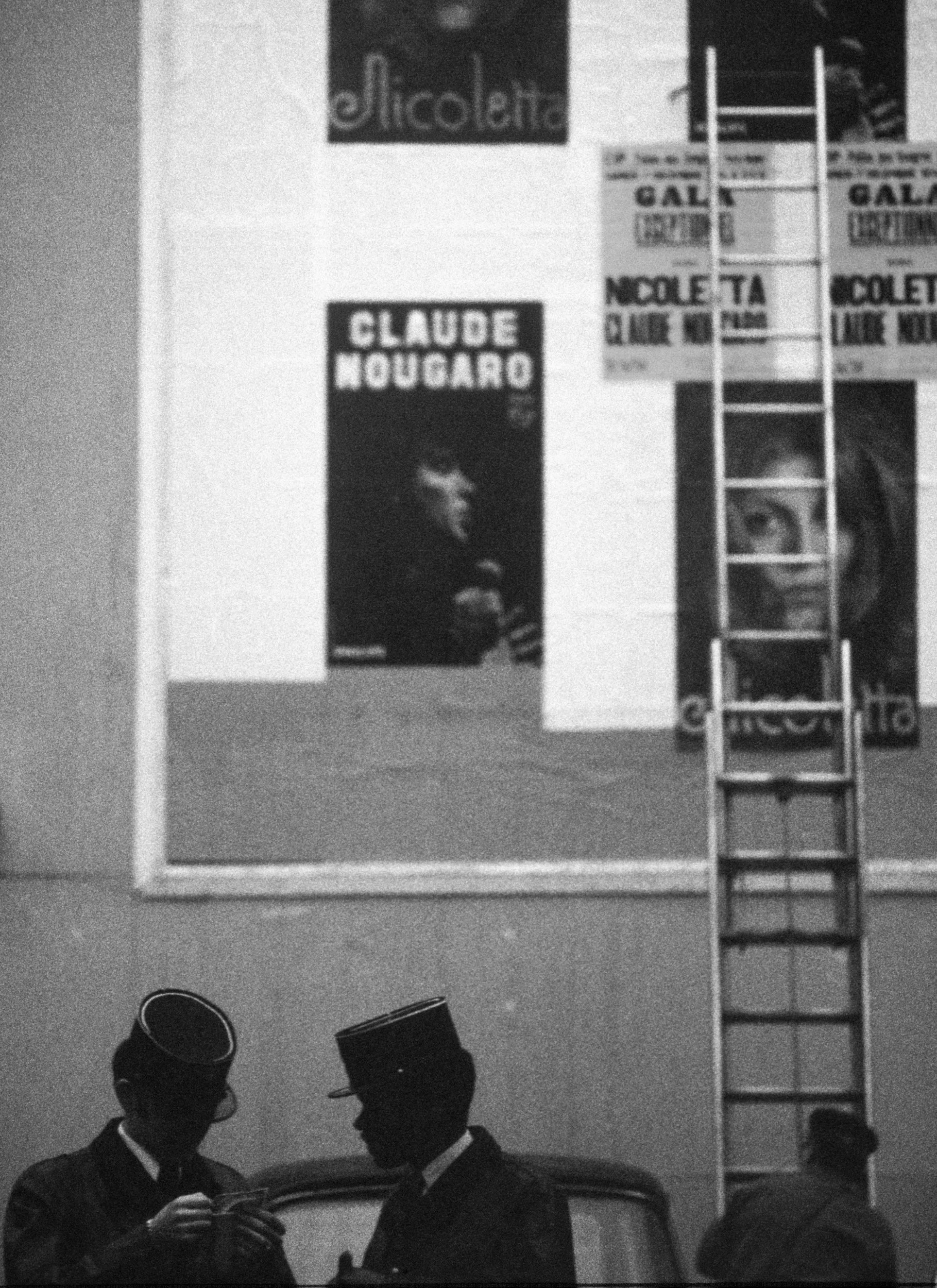 Black and white photo of two men in military uniforms and hats talking, with posters of Claude Nougaro and other events on the wall behind them, and a ladder leaning against the wall.