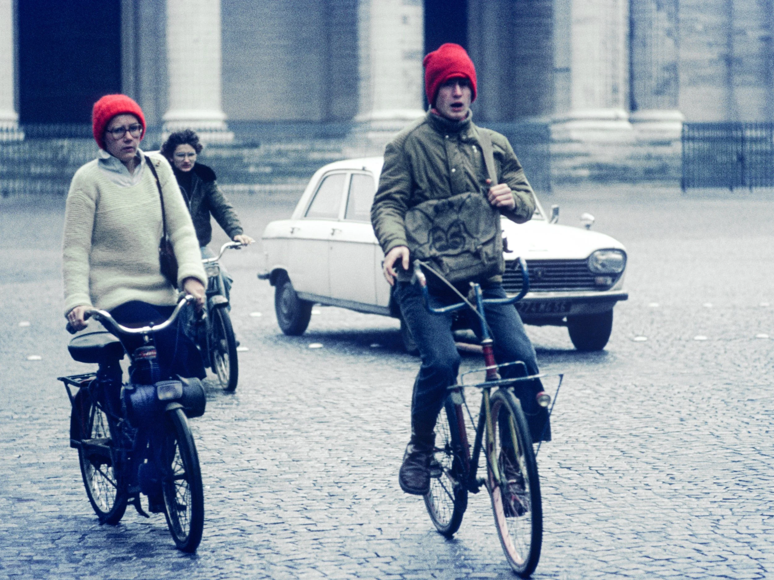 Three children, two girls and one boy, riding bicycles on a cobblestone street in cold weather, all wearing red beanies. A vintage car is parked in the background near historic stone and columned building.