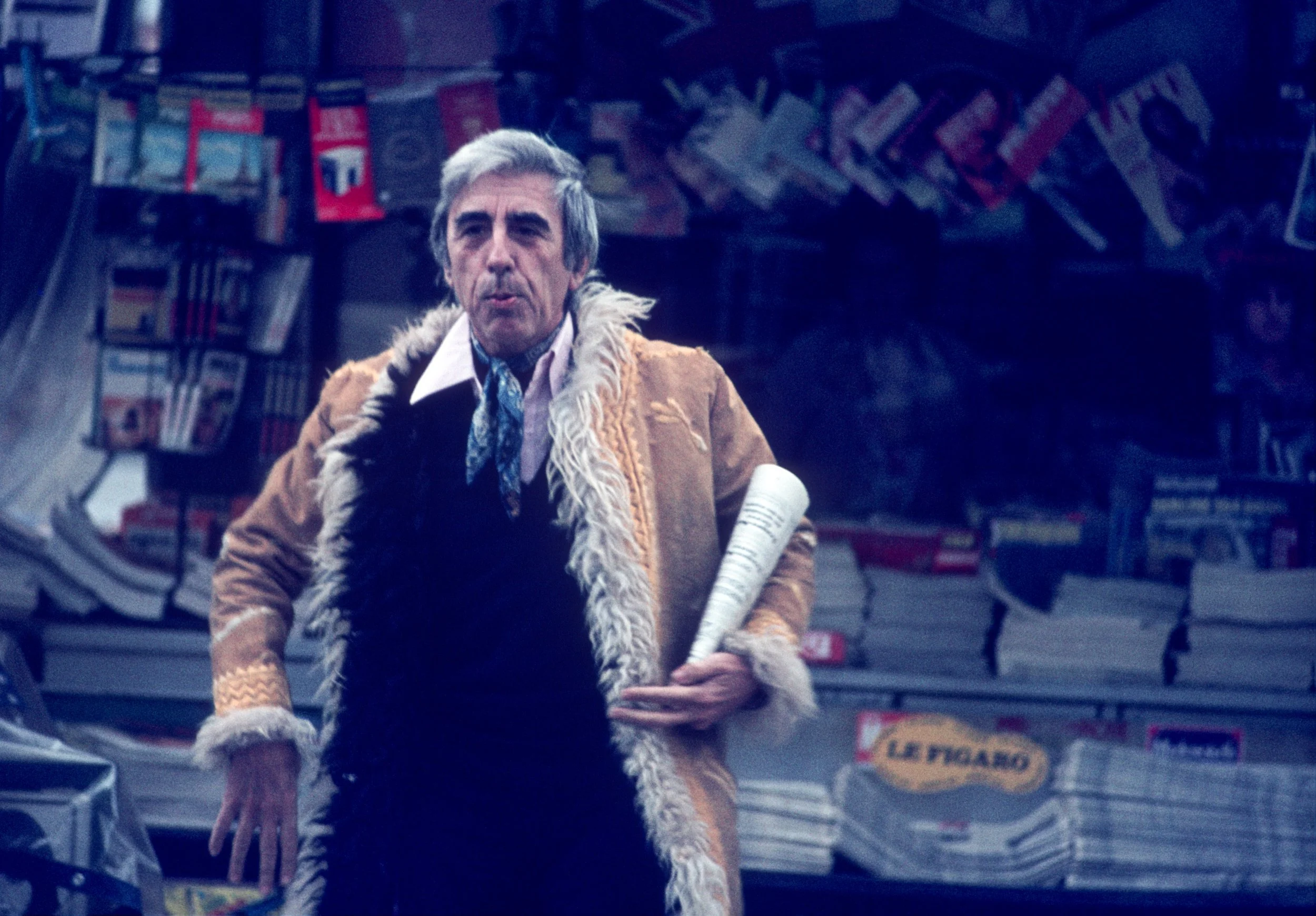 A man with gray hair and light skin wearing a costume with a brown fur-lined coat, a white shirt, and a blue dicky standing in front of a newsstand with magazines and newspapers, holding a rolled-up newspaper.