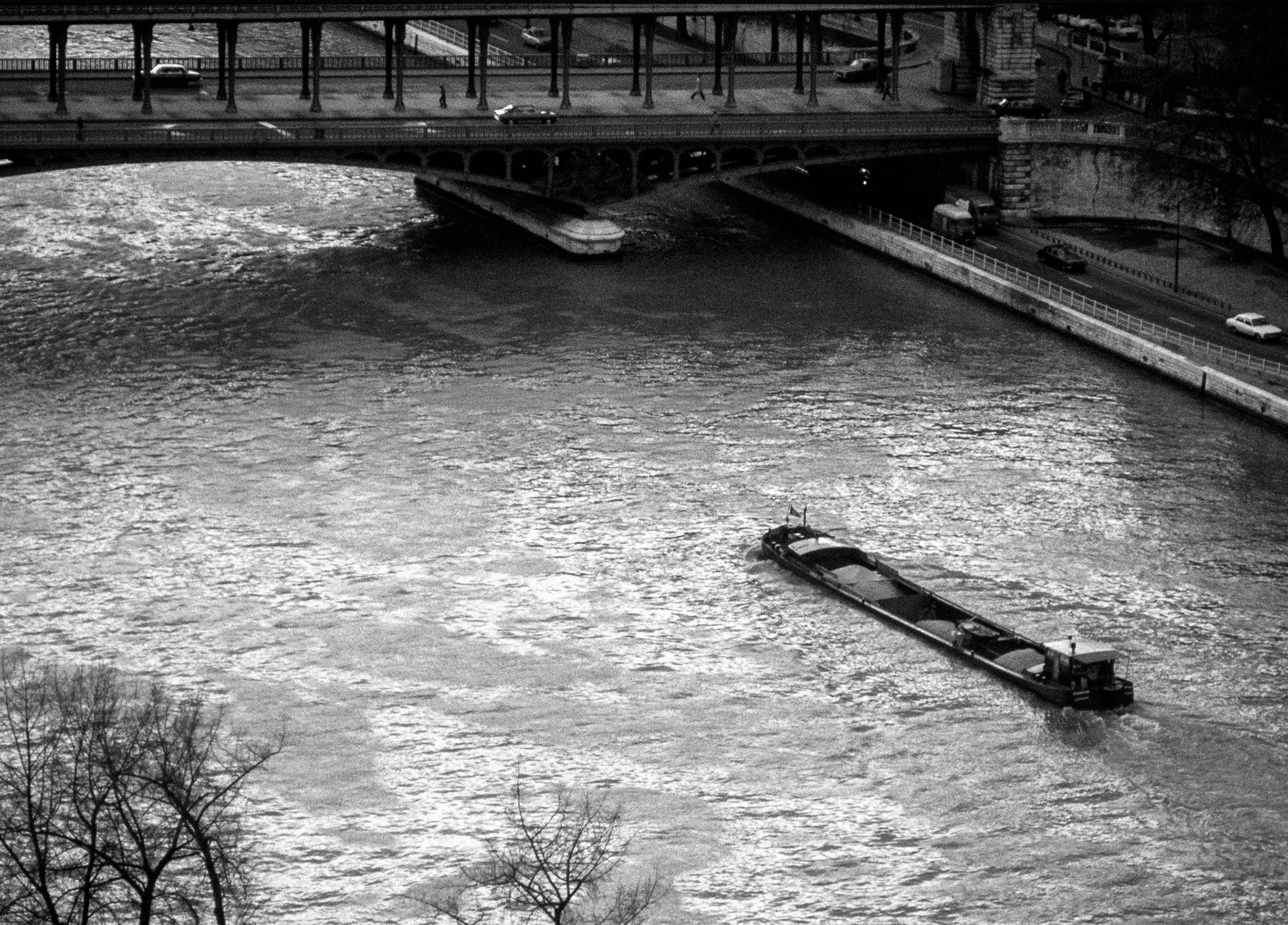 A black and white photograph of a river with a boat moving through the water. There is a bridge crossing over the river with cars traveling on it, and a road running parallel under the bridge. Bare trees are visible at the bottom of the image.