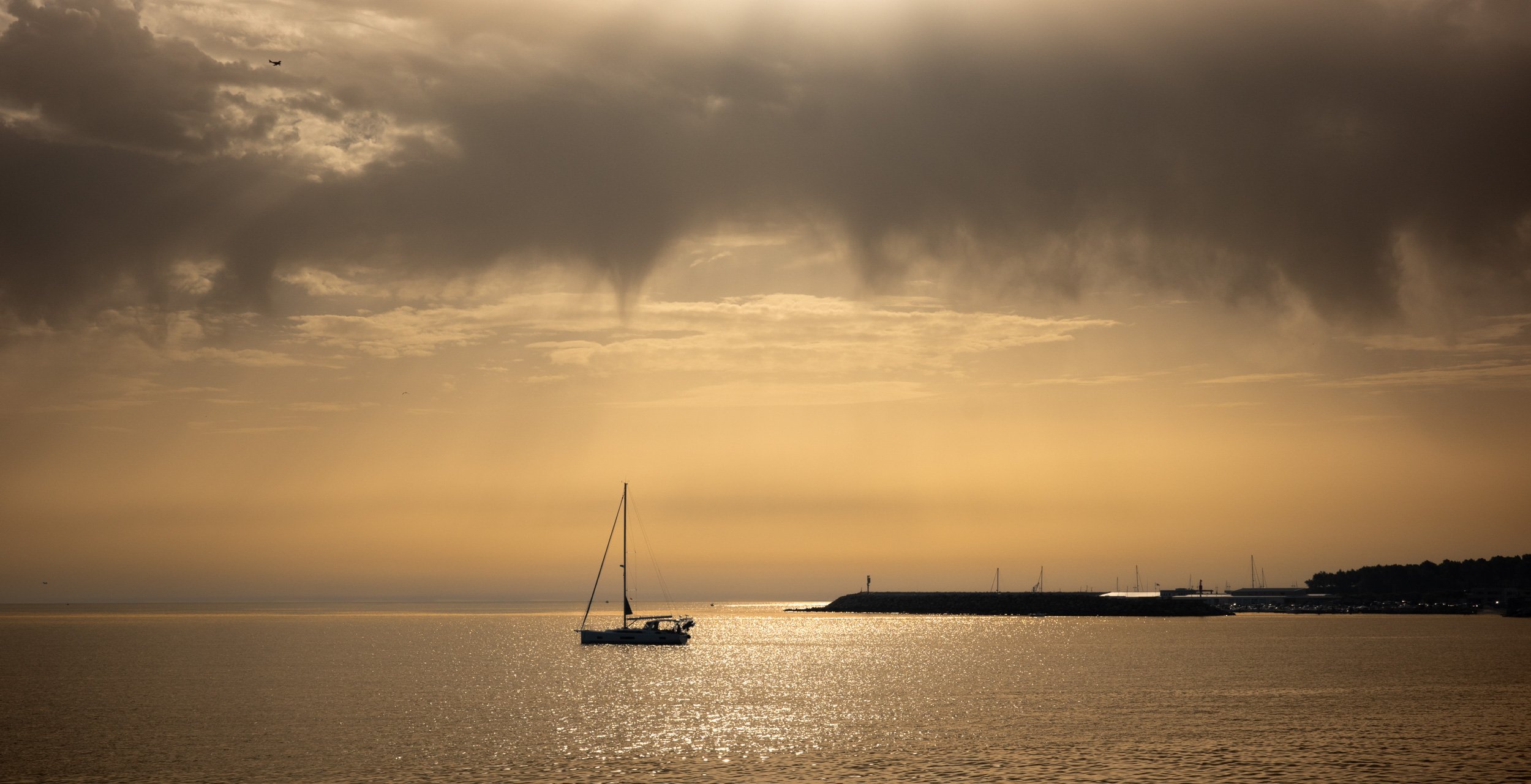 Sunset over a calm body of water with a single sailboat in the foreground. Dark clouds are present in the sky with a small airplane flying overhead.