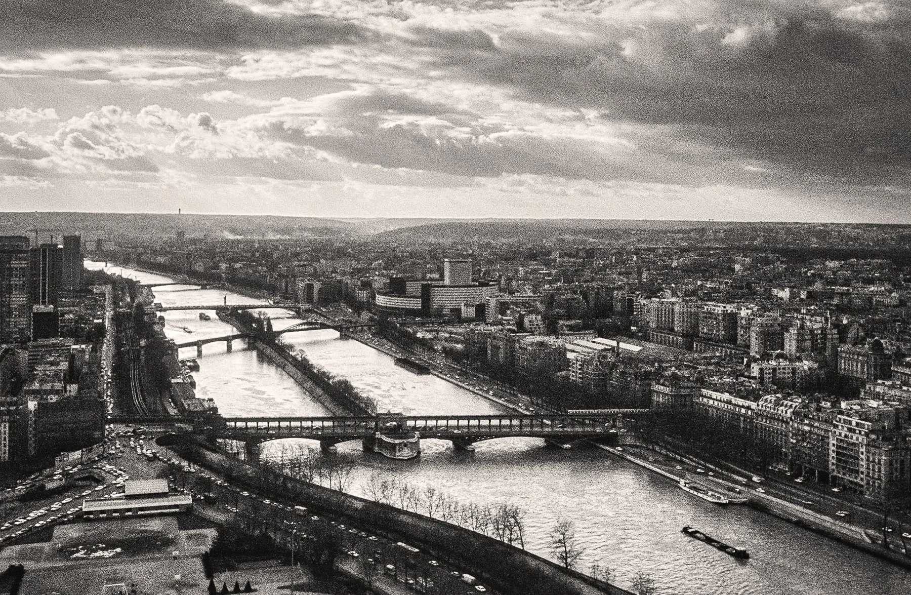A black and white aerial view of a cityscape with a river flowing through the center, multiple bridges crossing the river, and a mix of high-rise and lower buildings. The sky has scattered clouds.