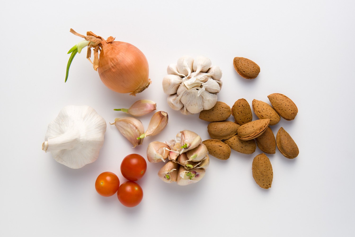 A collection of vegetables and nuts on a white surface, including an onion with green sprouts, garlic cloves, garlic bulbs, cherry tomatoes, garlic, and almonds.